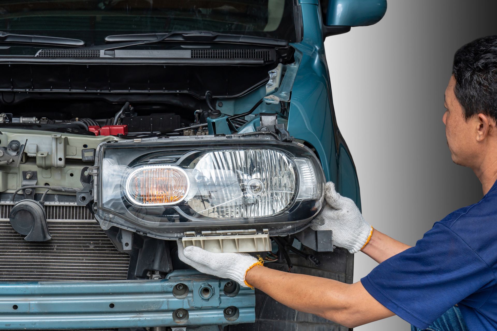 Mechanic removing a car headlight, wearing gloves, in front of a blue vehicle.
