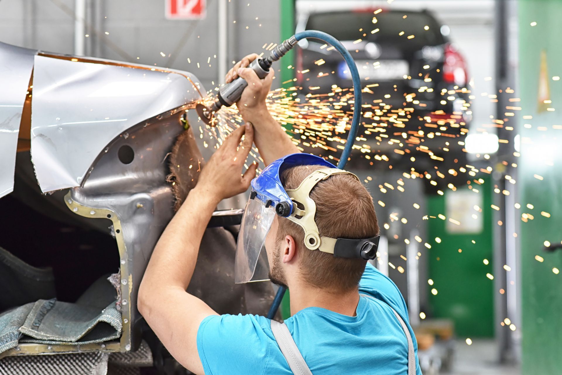 A mechanic grinds metal on a car, creating sparks. They wear a face shield in a garage.