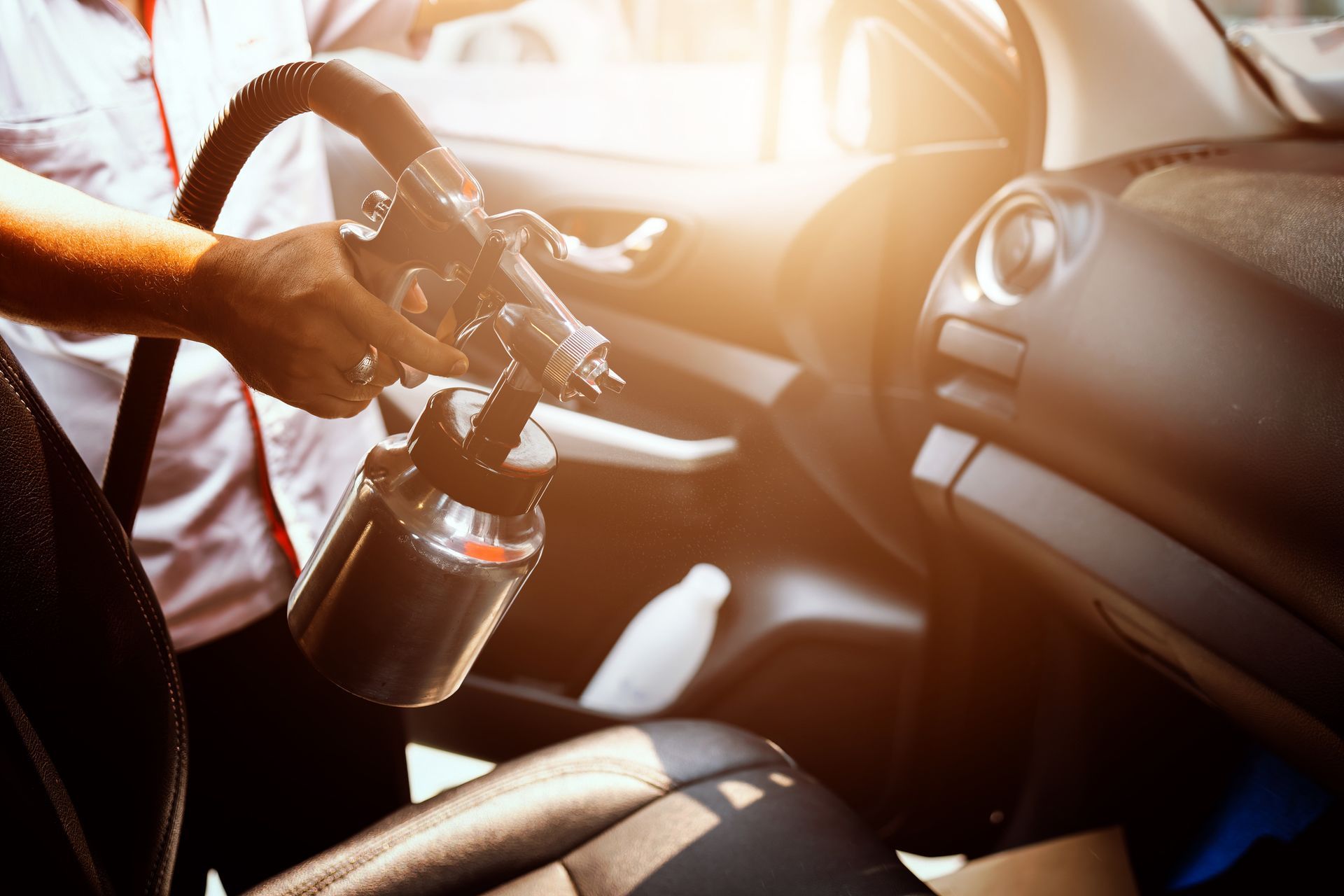 Person sprays car interior with a cleaning gun; sunlight streams through the window.