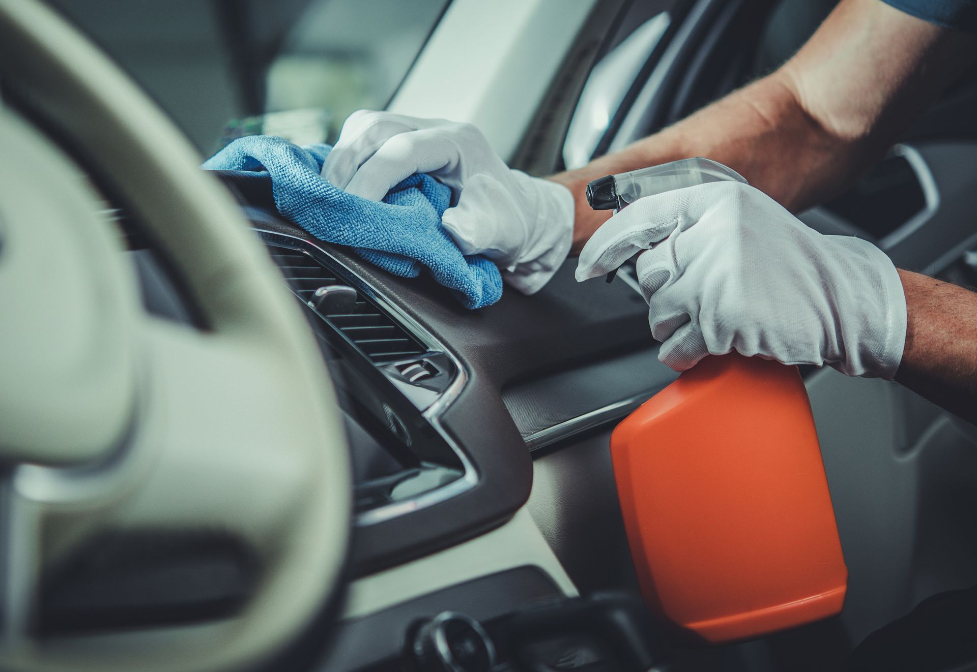 Person wearing gloves cleaning car dashboard with spray bottle and blue cloth.