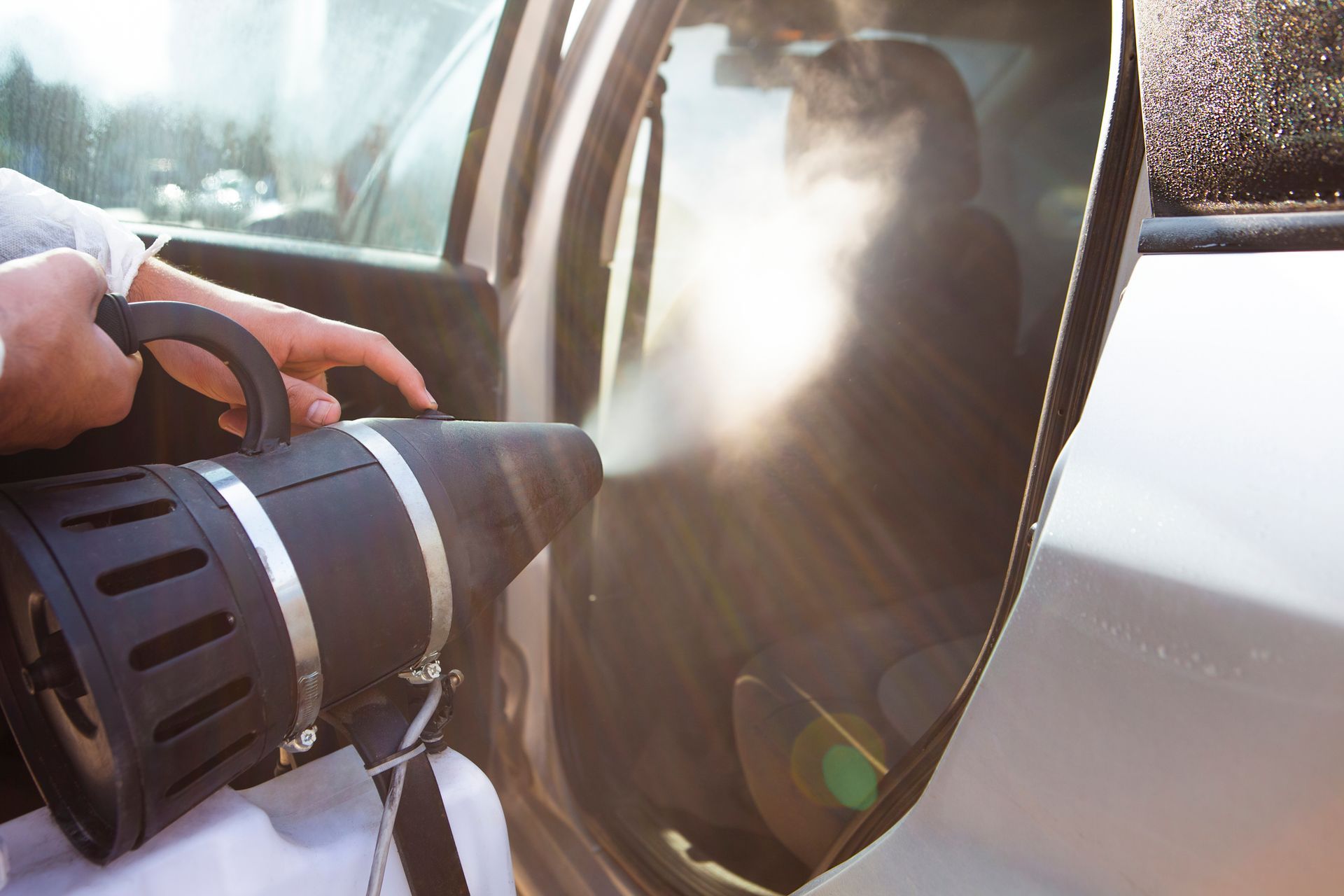 Person disinfecting a car interior with a fogging machine, emitting a white mist.