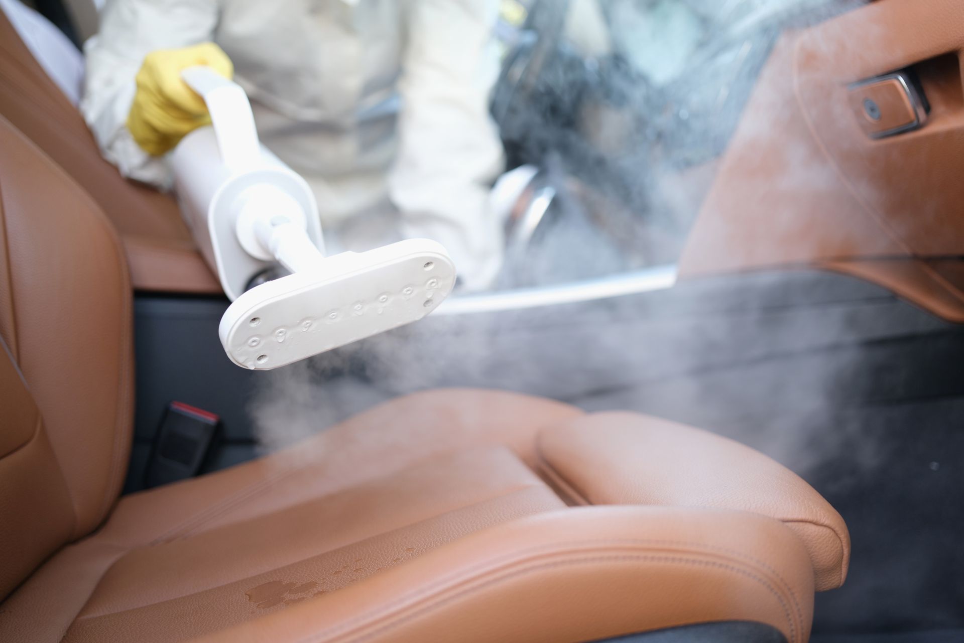 Person in protective gear steam cleaning a brown car seat.