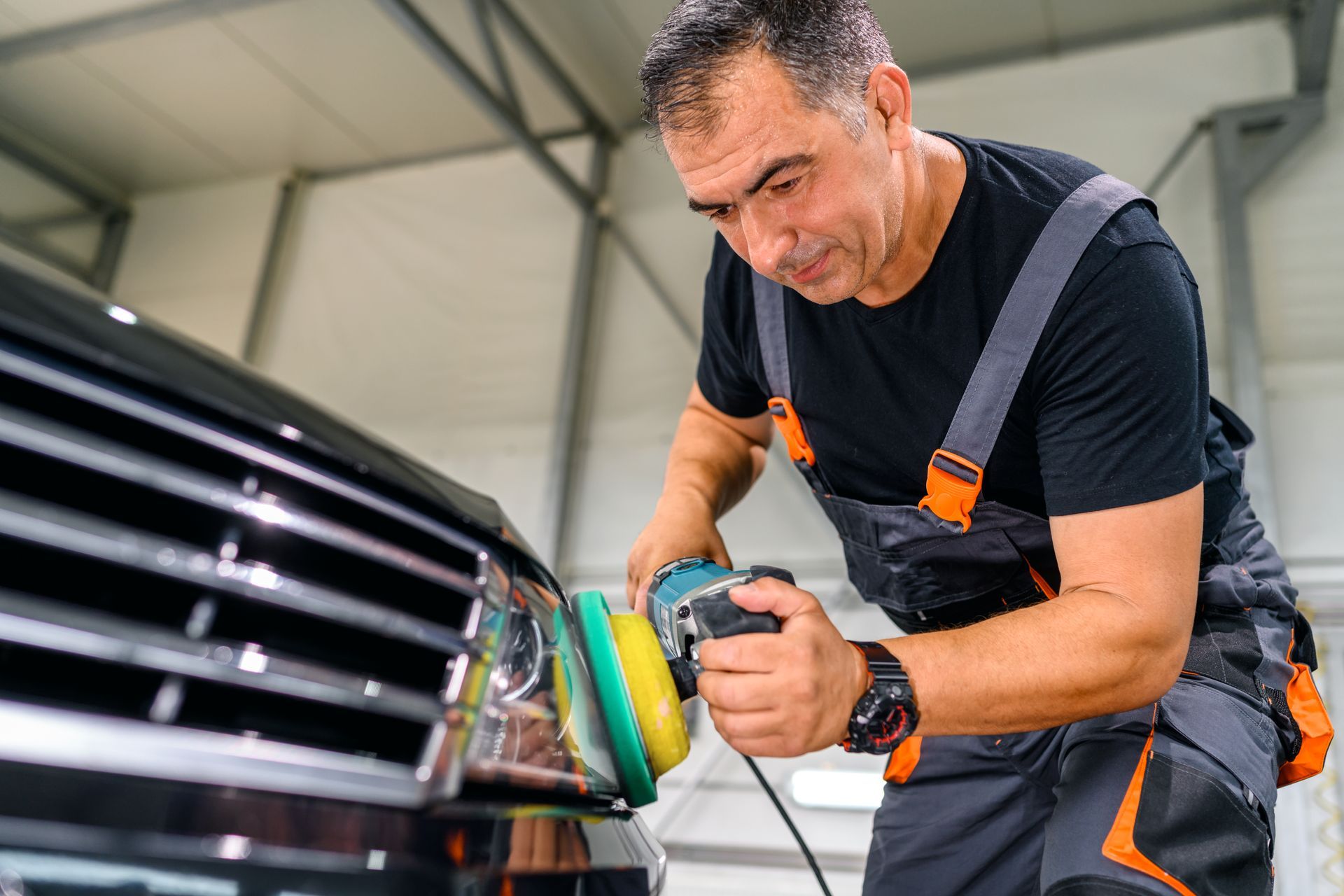 Man polishing a black car with a power buffer in a garage.