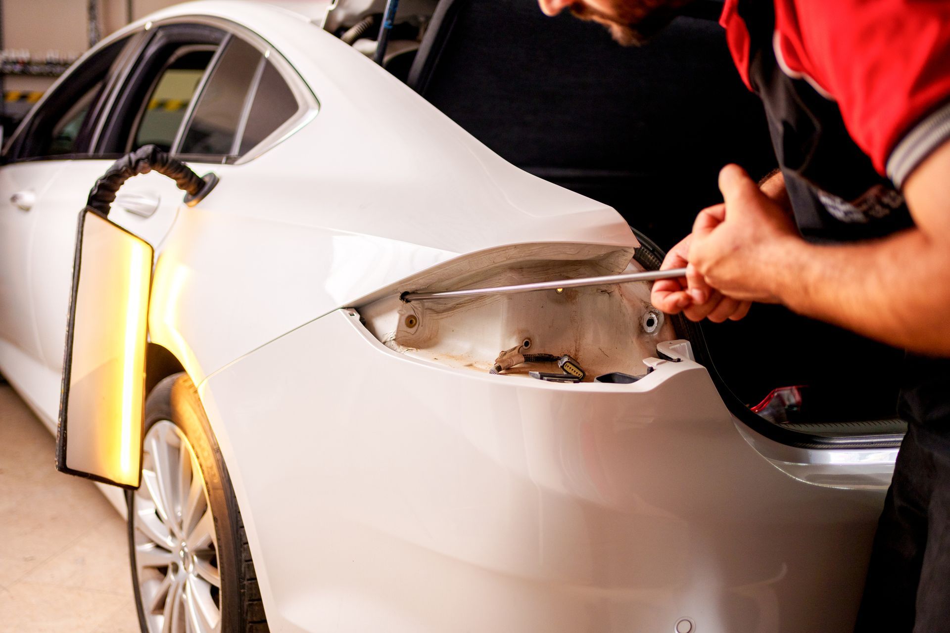 Mechanic repairing a white car's rear with a tool; a light highlights the damage.
