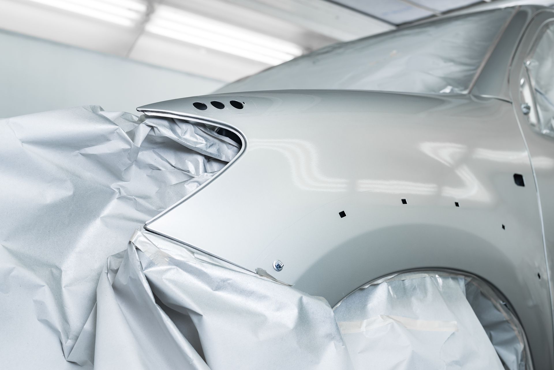 Car being prepped for paint job in a paint booth. Covered in protective sheeting; silver vehicle panels.