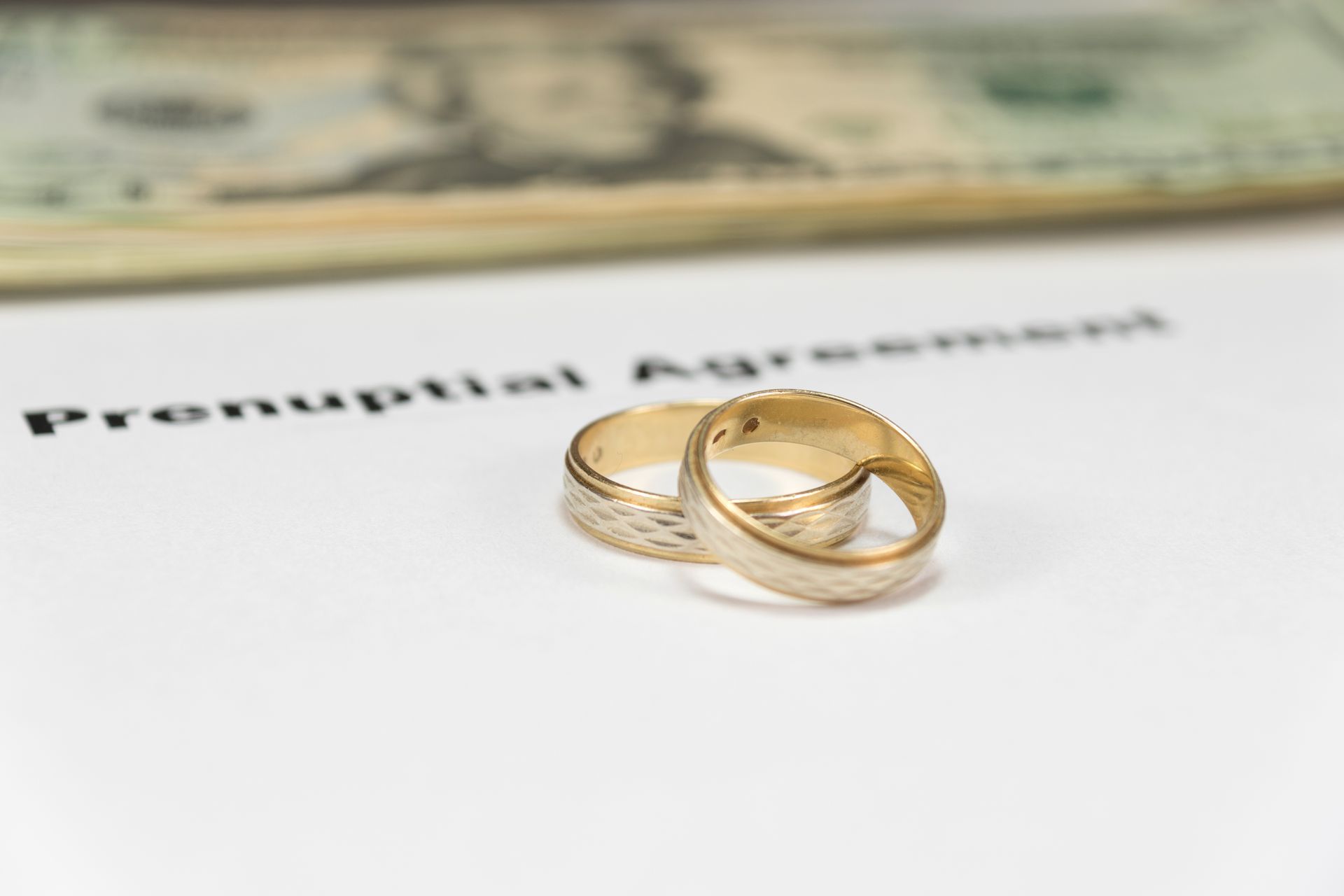 Two wedding rings on a prenup document, with money visible in the background.