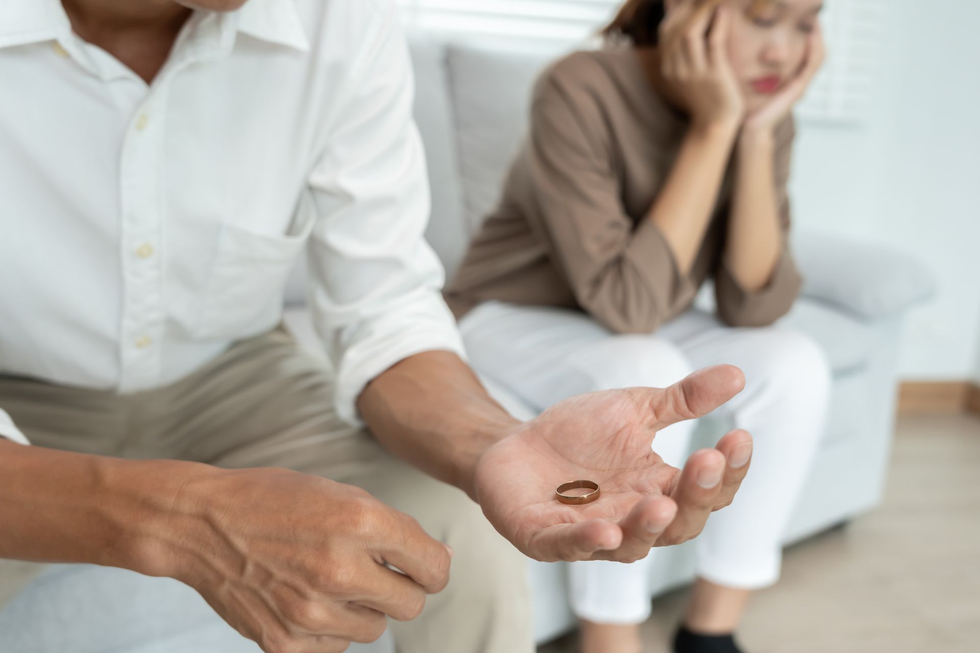 Man holds wedding ring, woman looks sad on sofa, indoor setting.