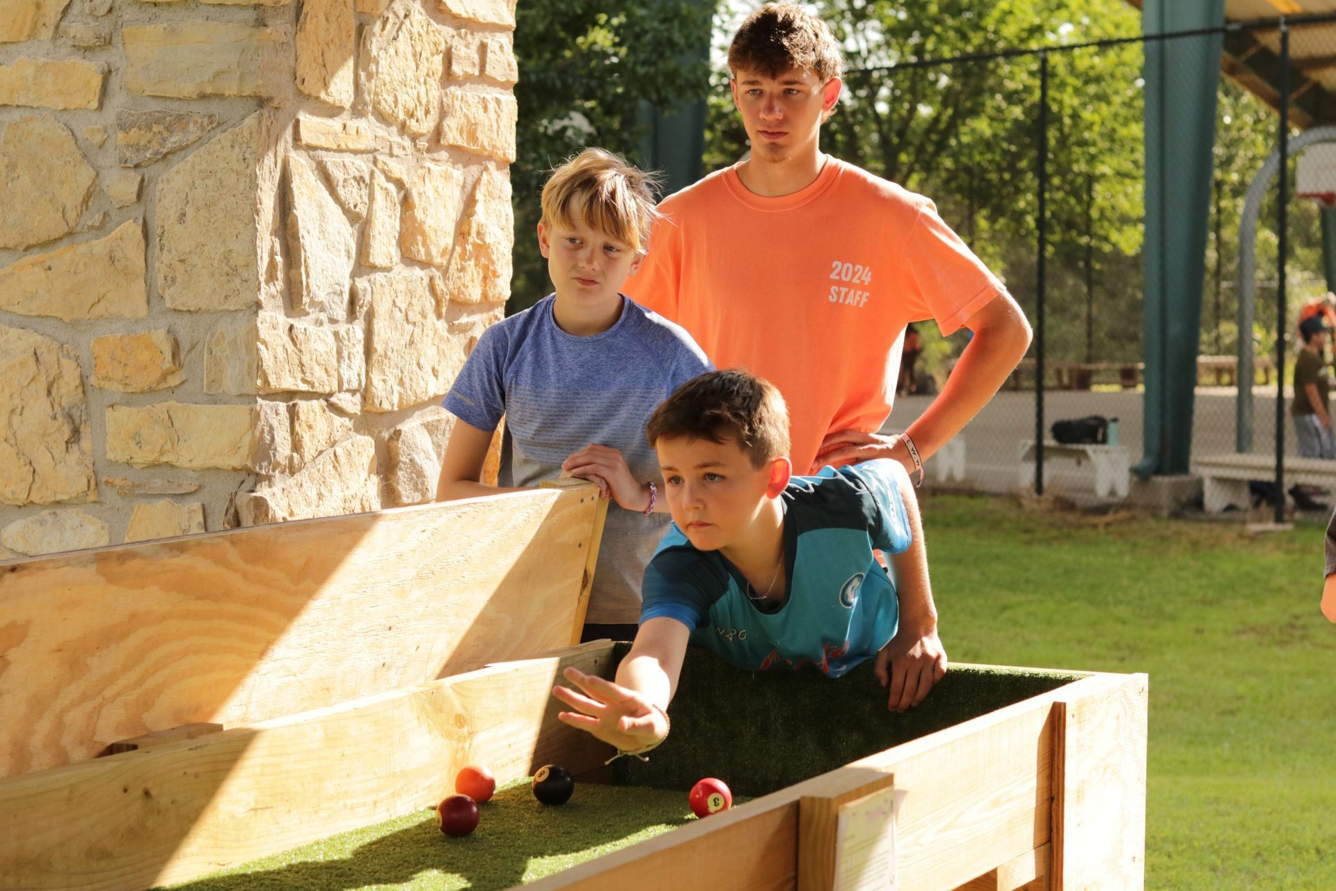 Three young boys are playing a game of bocce ball in a wooden box.