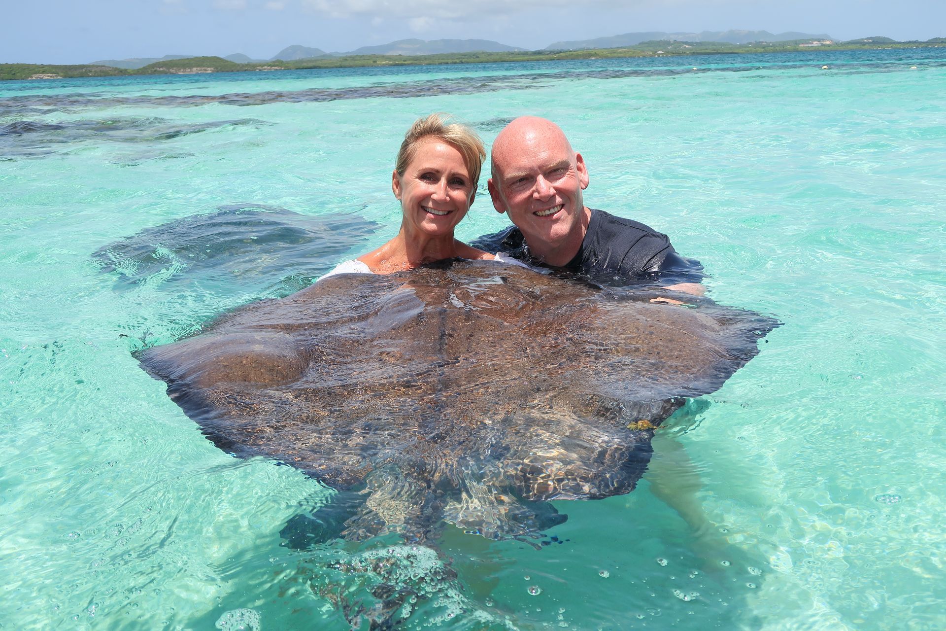 Couple in shallow turquoise water with a large stingray; smiling faces, sunny day.