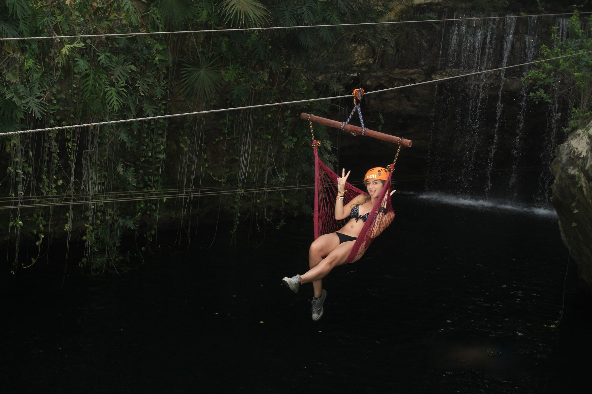 Woman in bikini on a hammock swing over dark water near a waterfall, giving a peace sign.