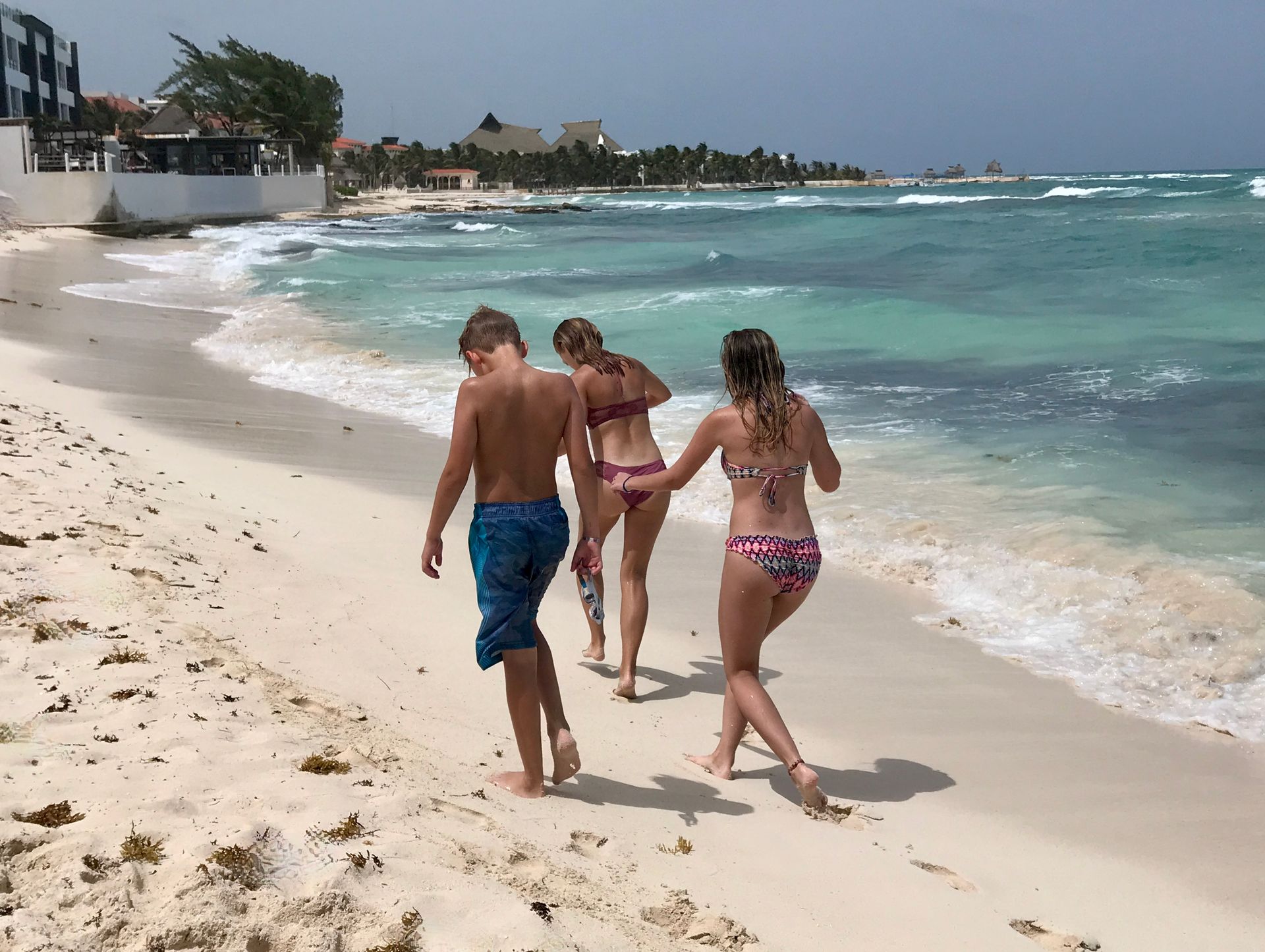 Three children walking on a beach: two teenagers in bikinis and a shirtless boy in blue swim trunks.