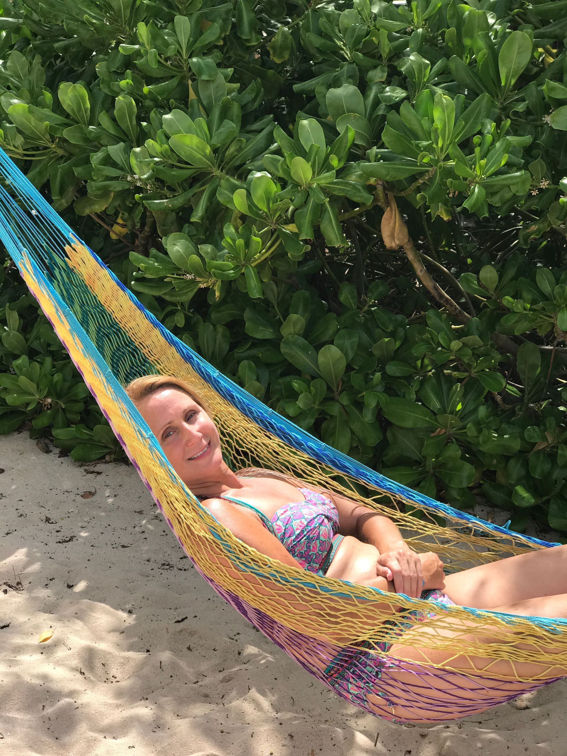 Woman smiling in a colorful hammock on a beach, with green foliage in the background.