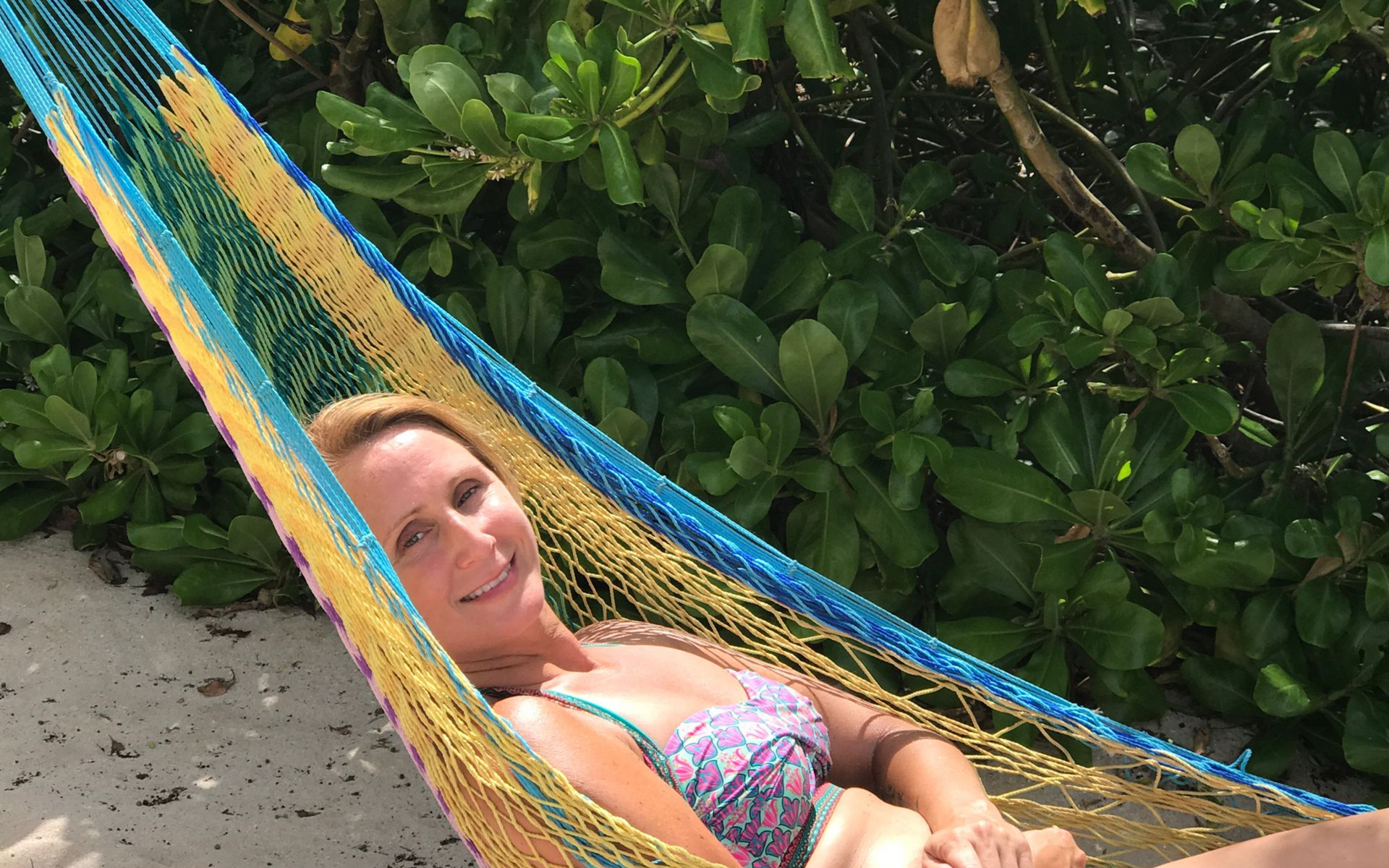 Woman in swimsuit relaxes in colorful hammock near green foliage and sand.