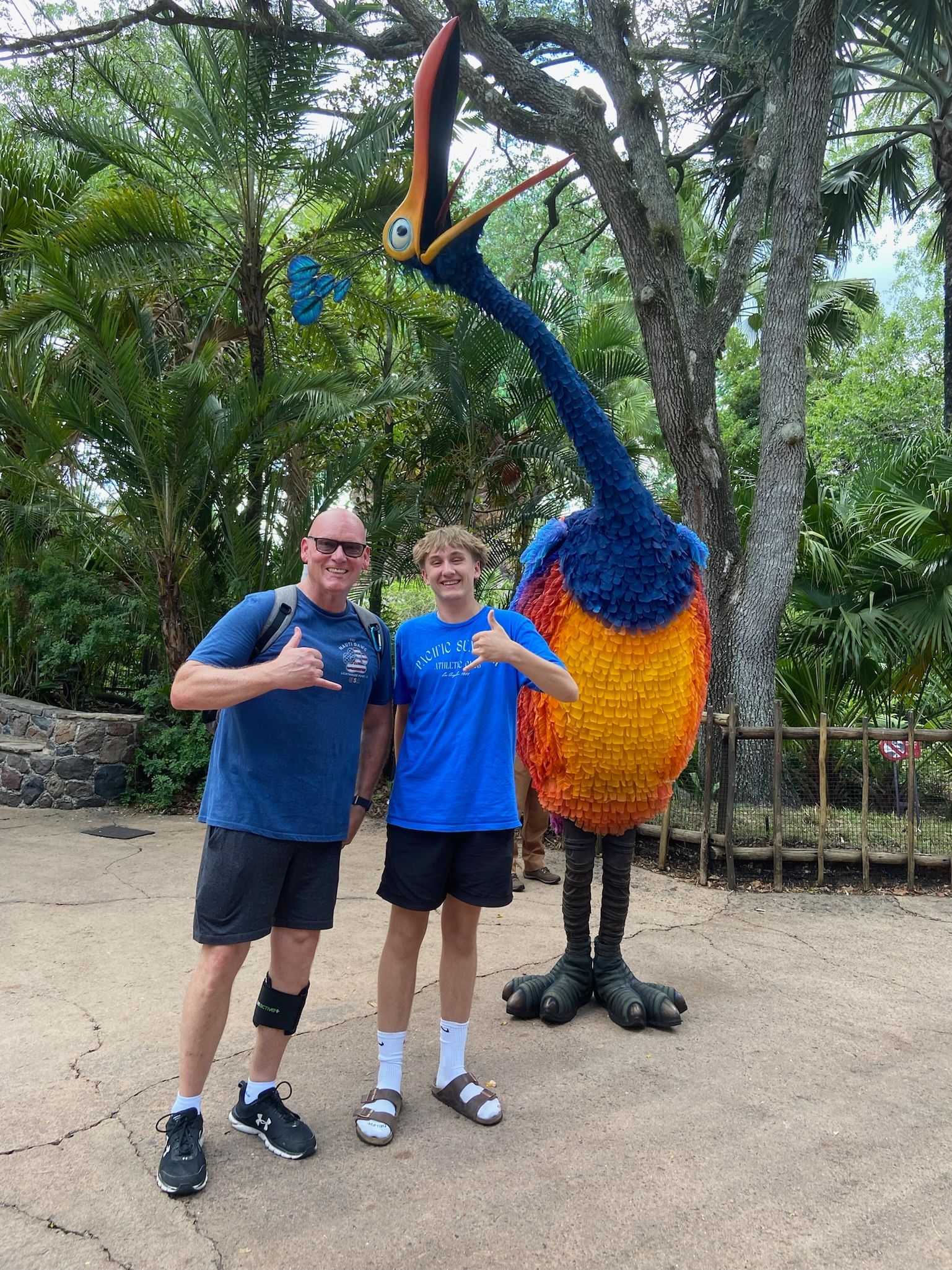 Man and boy pose with a giant Kevin bird statue at an outdoor park; both make shaka hand gestures.