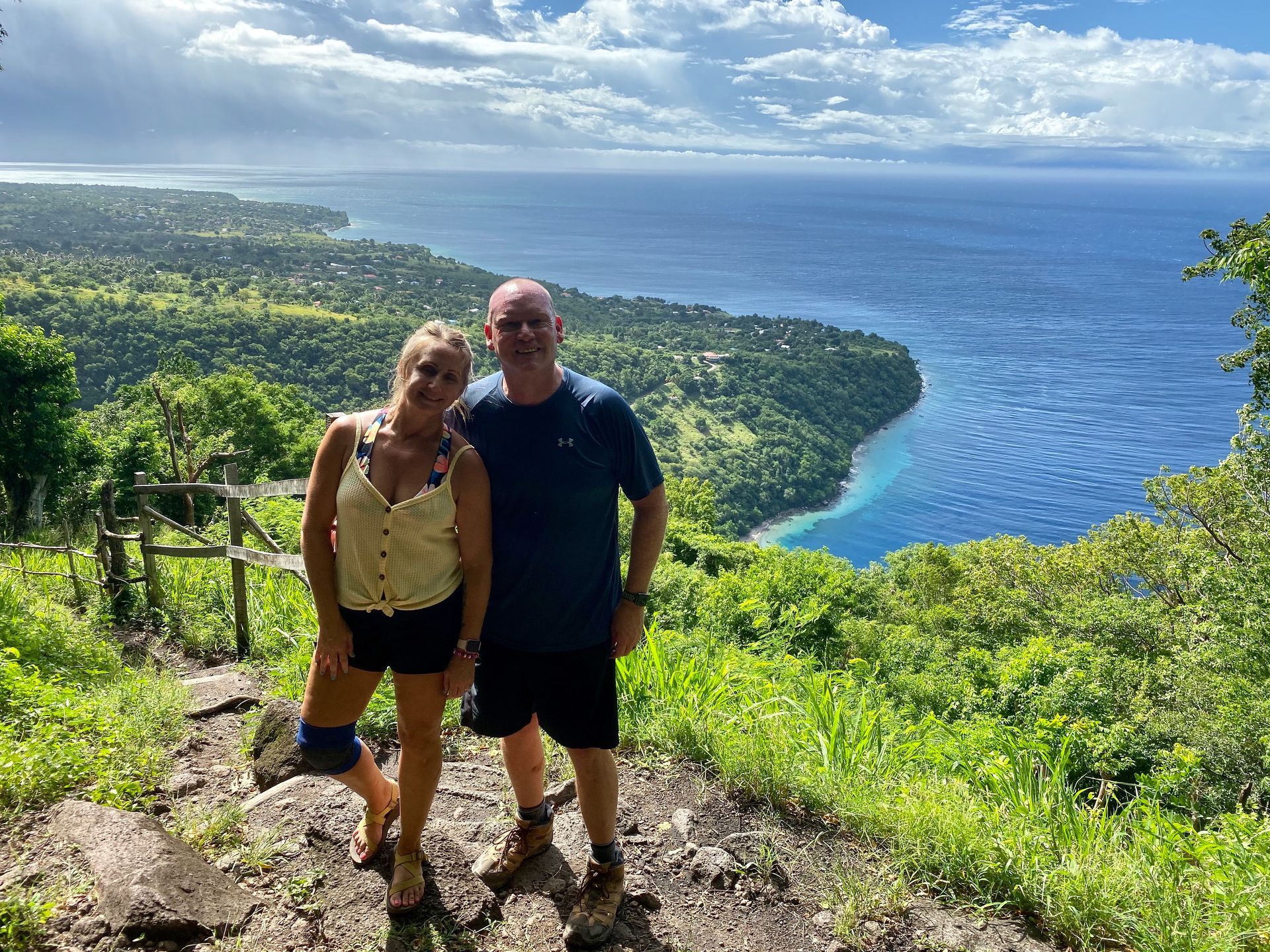 Two people standing on a lush hillside overlooking a blue bay and coastline under a cloudy sky