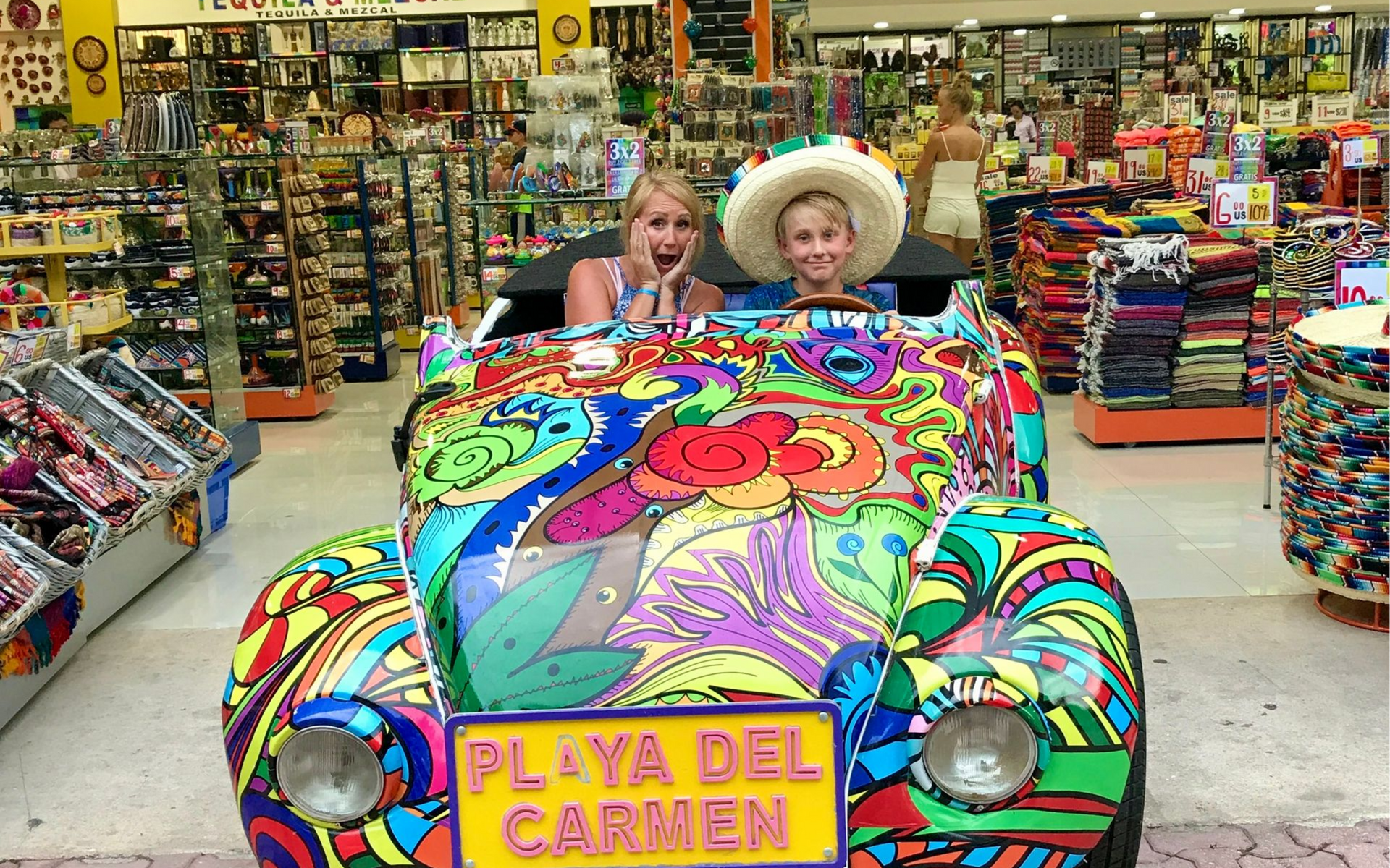 Two people posing in a colorful Volkswagen cutout in a Playa del Carmen souvenir shop. One wears a sombrero, both smile.