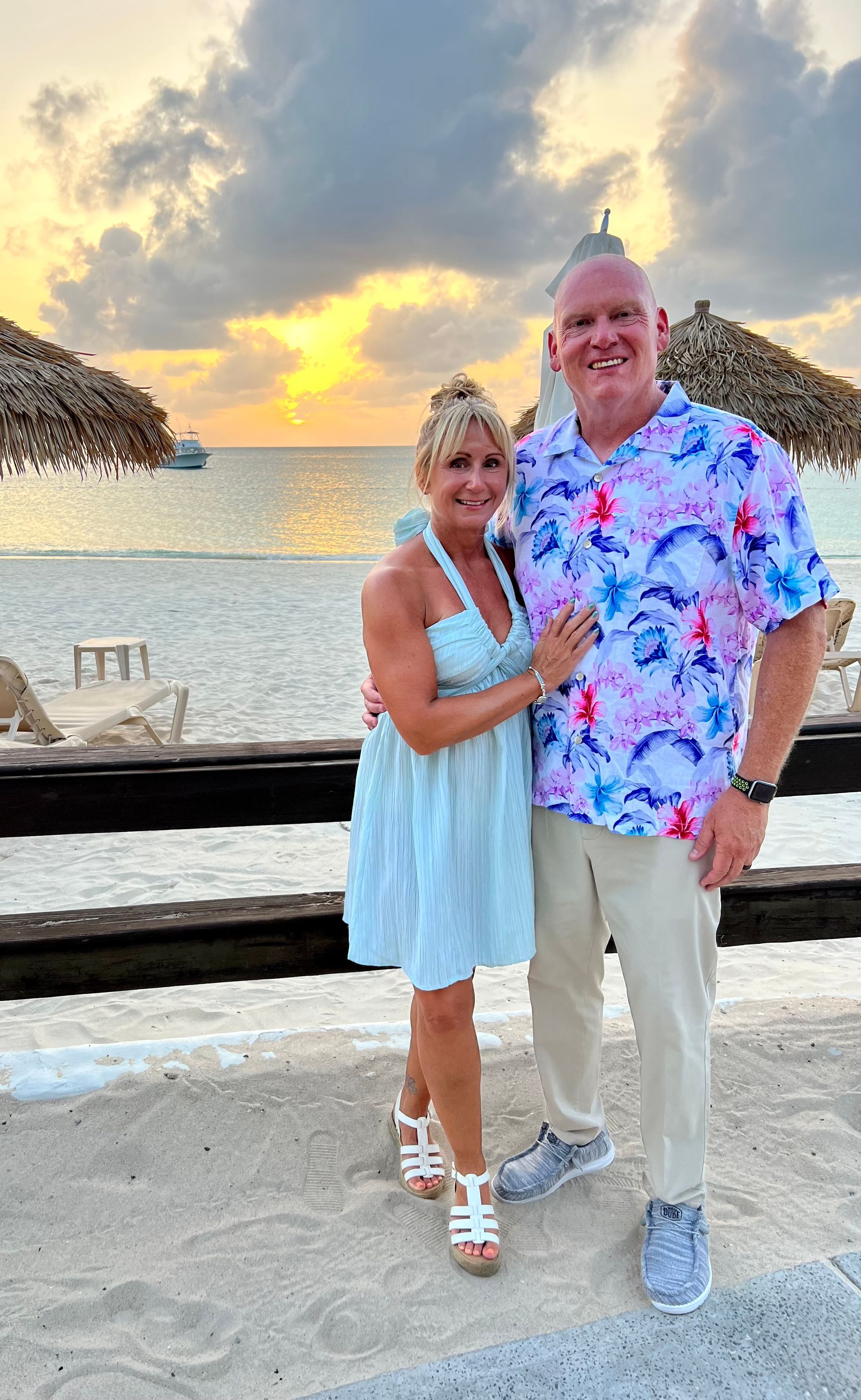 Couple smiles on beach at sunset. Woman in blue dress, man in floral shirt, sand, water.