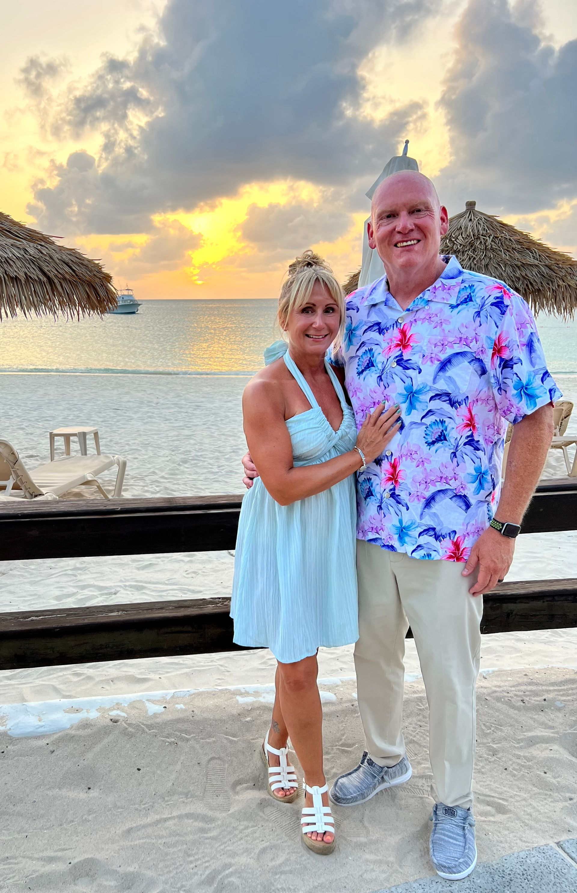 Couple at a beach sunset. Woman in light blue dress, man in Hawaiian shirt. Beach, sea, and thatched roofs visible.