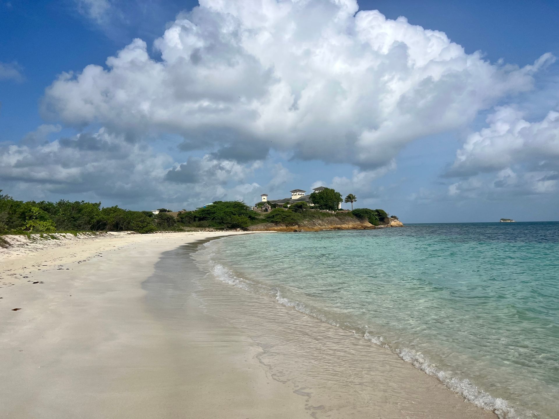 Sandy beach with clear turquoise water, lush green vegetation, and a cloudy blue sky.