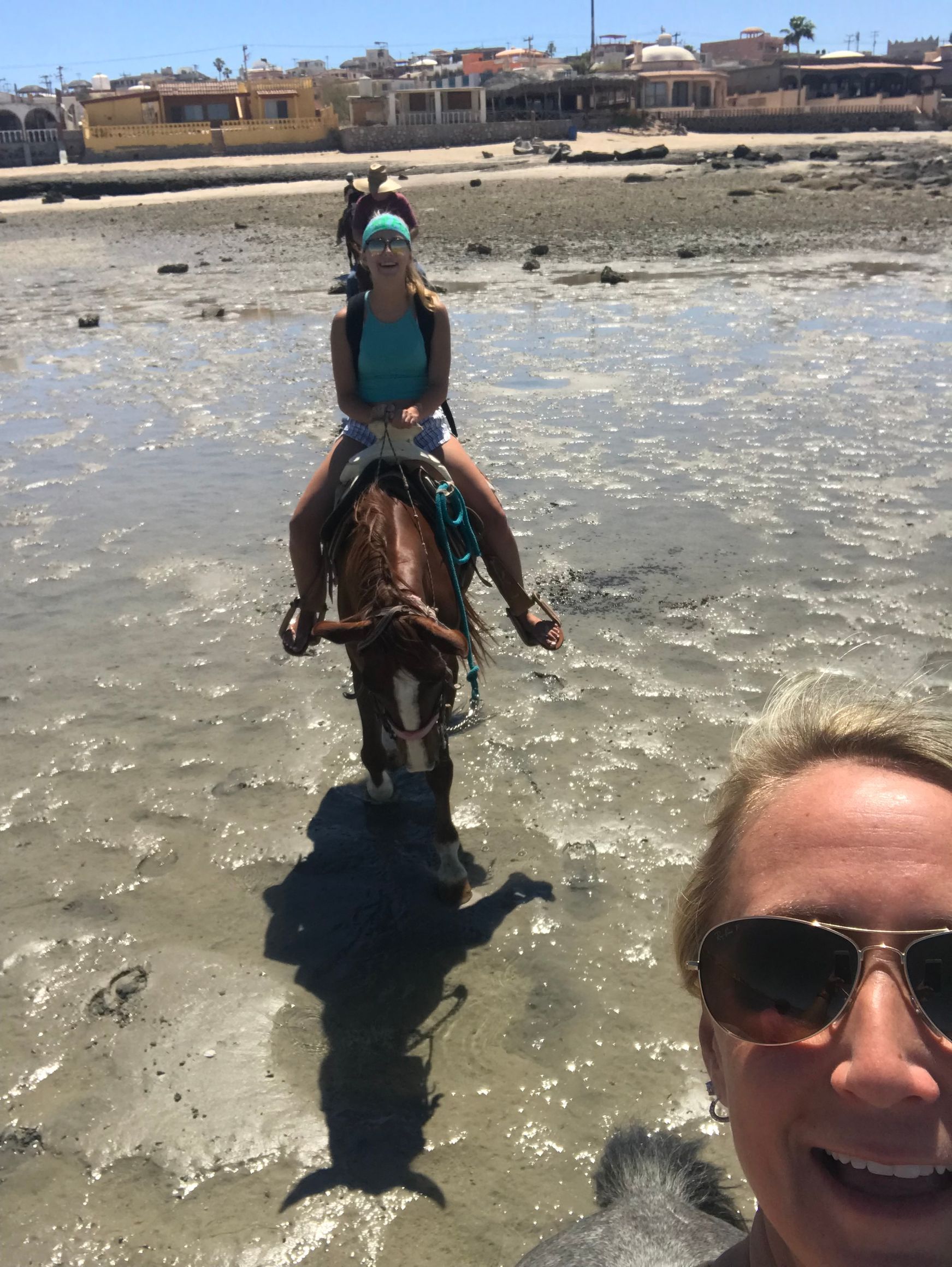 Woman riding a horse in shallow water; another woman takes a selfie. Sunny day, beach setting.