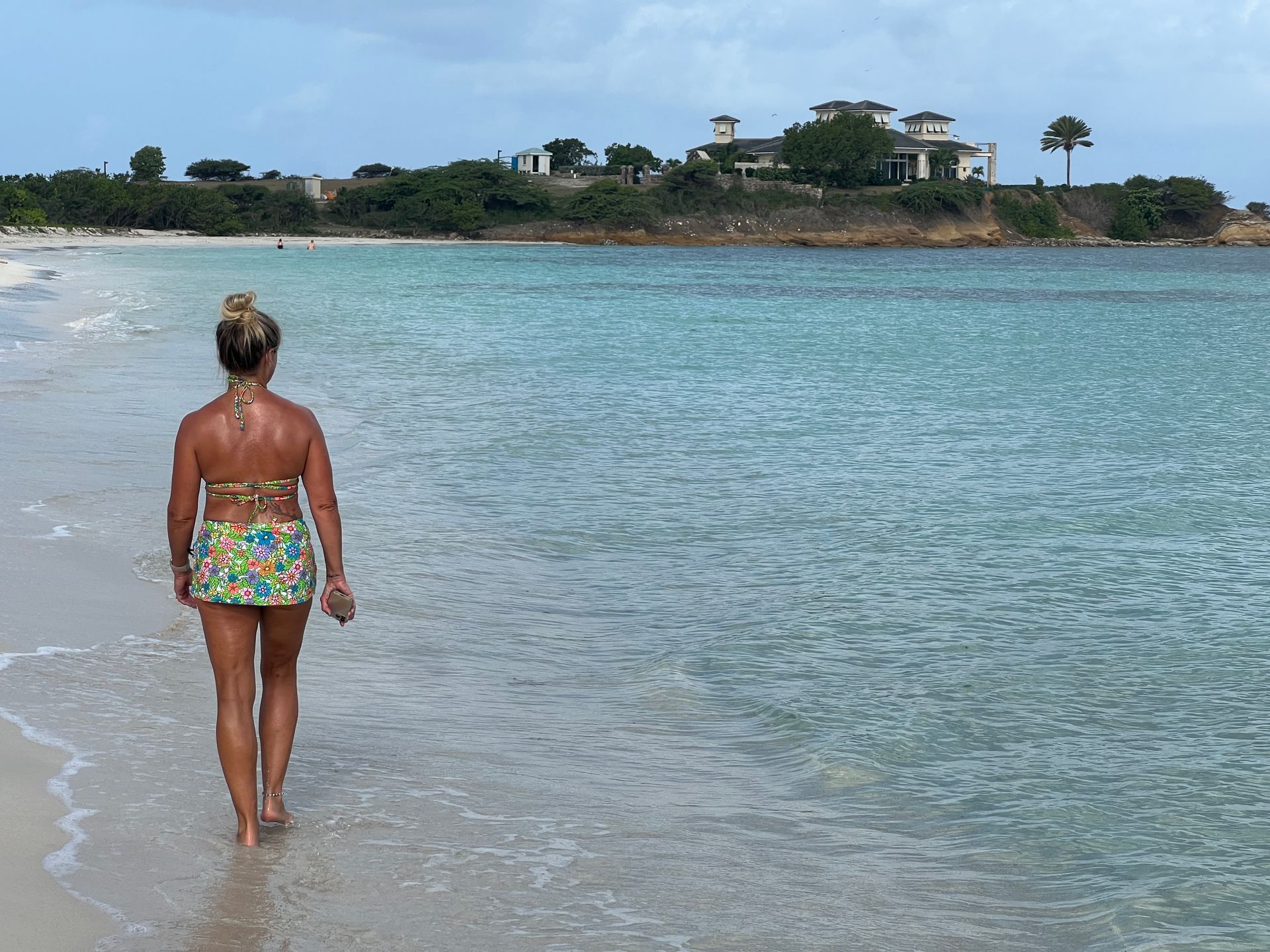 Woman in floral bikini walking on beach, turquoise water, buildings on shore.