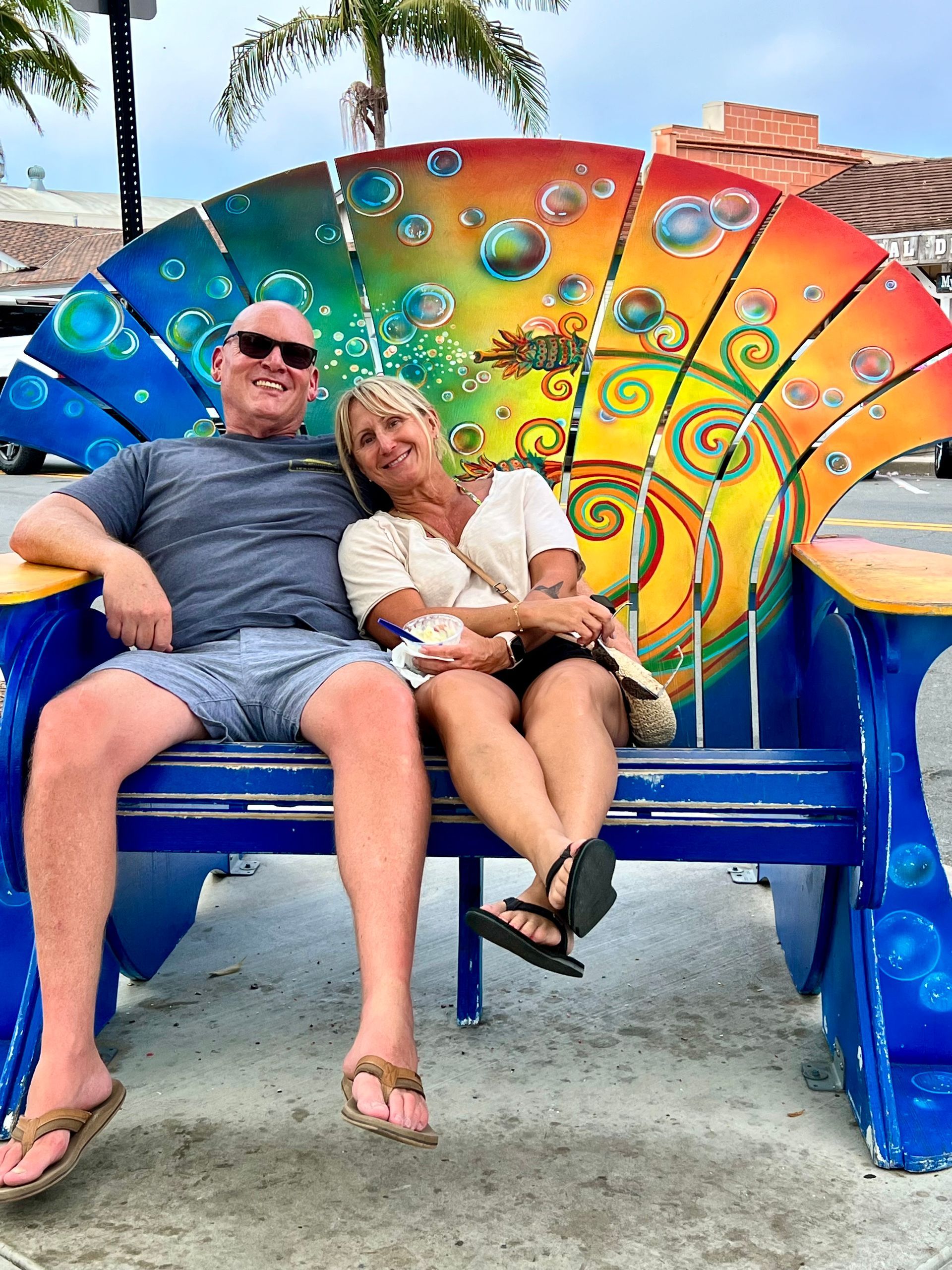 Couple sitting on a colorful, seashell-shaped bench in Carlsbad, CA. Man wears sunglasses. Woman rests on his shoulder.