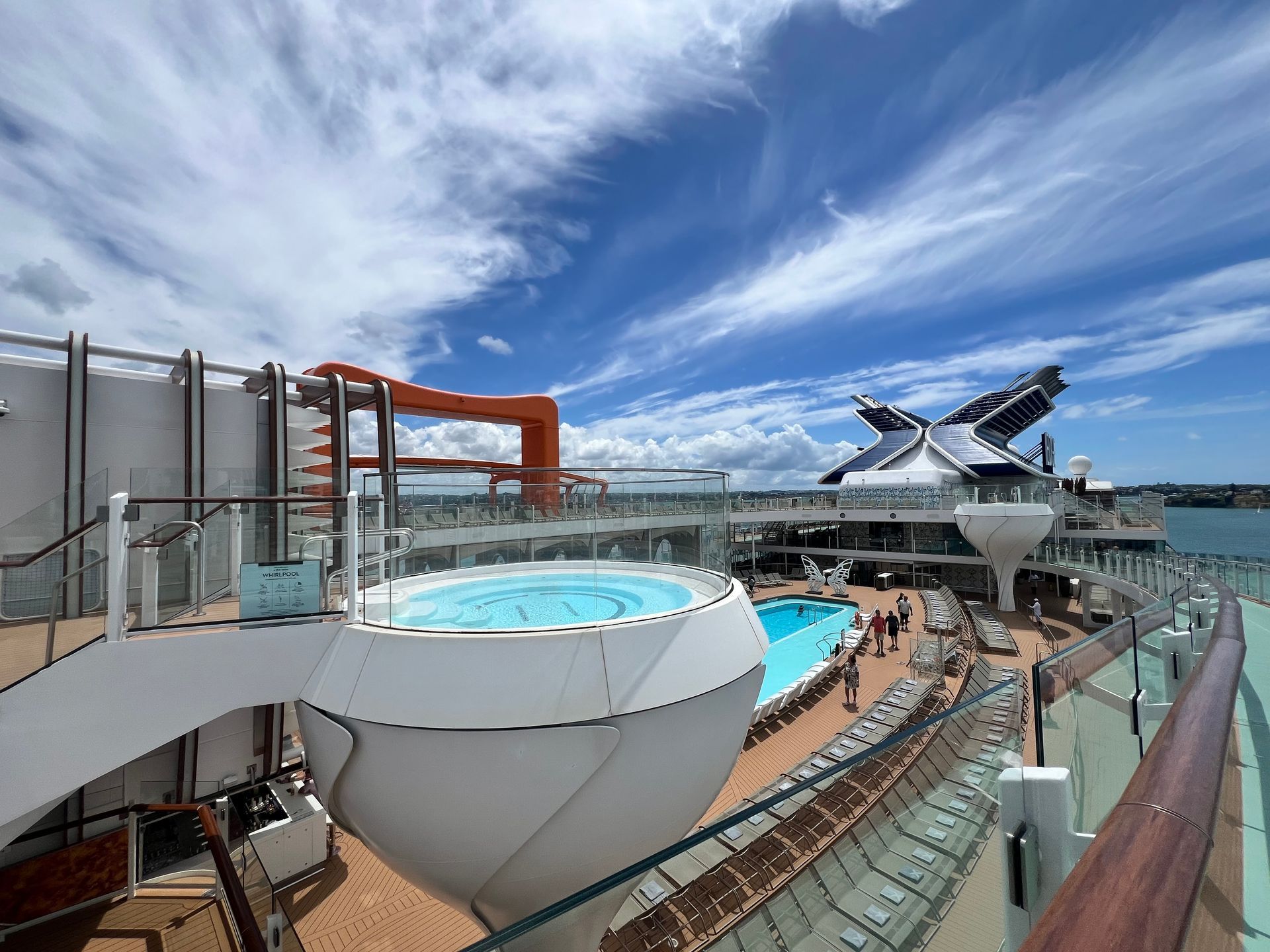 Cruise ship deck with pool, hot tub, and water slide under a blue sky with clouds.