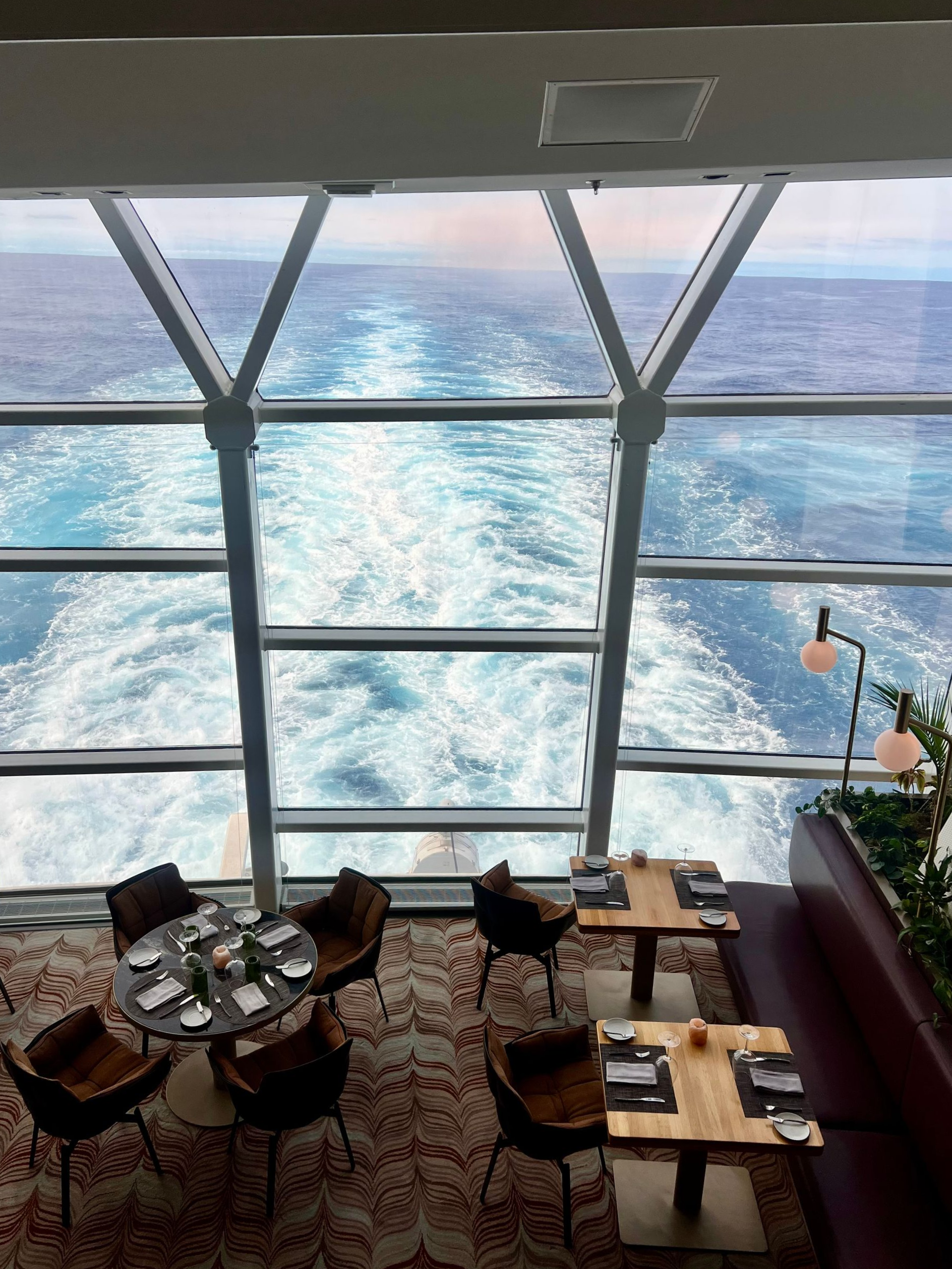 Restaurant with ocean view: Tables and chairs, ship's wake visible through geometric windows.