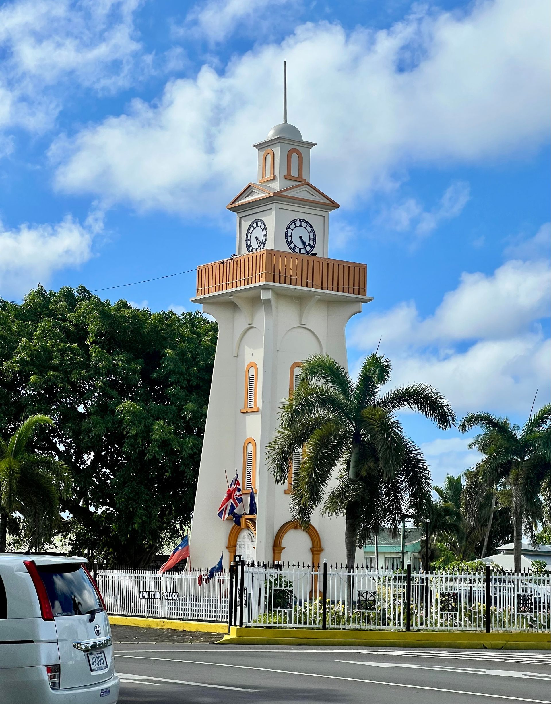 Clock tower in a park, white with clock faces, palm trees, and blue sky.