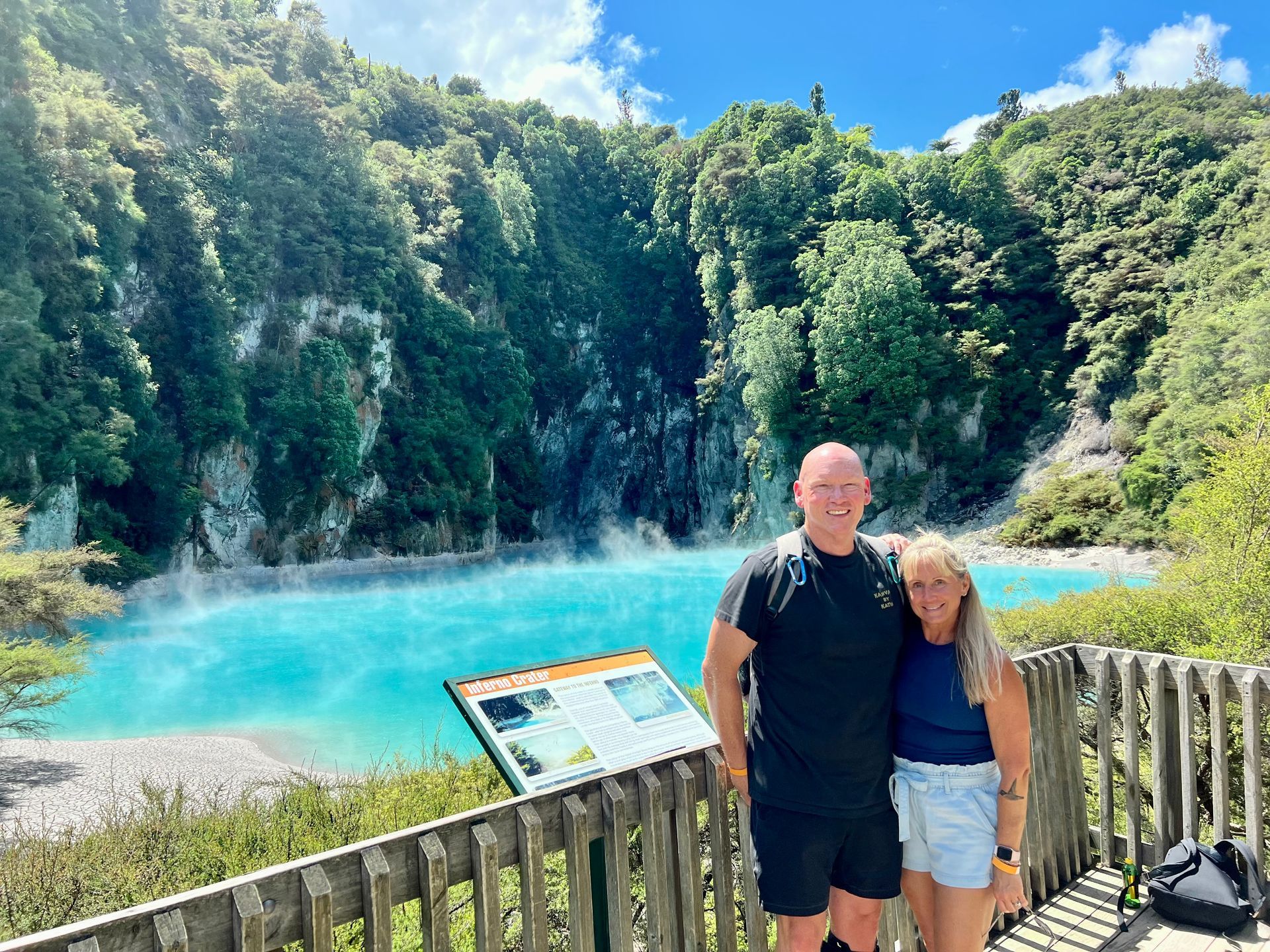 Couple poses by a turquoise lake in a geothermal area; signage and wooden railing in the foreground.