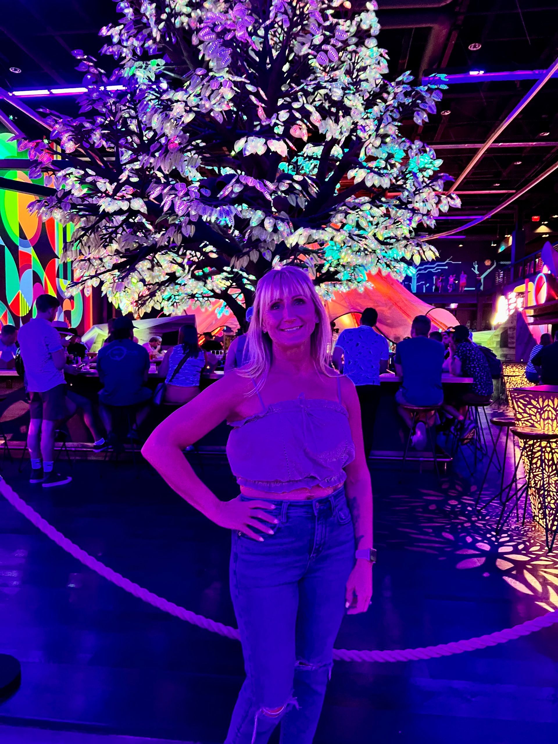 Woman in a bar in Las Vegas under a glowing tree, wearing jeans. Neon lights illuminate the space.