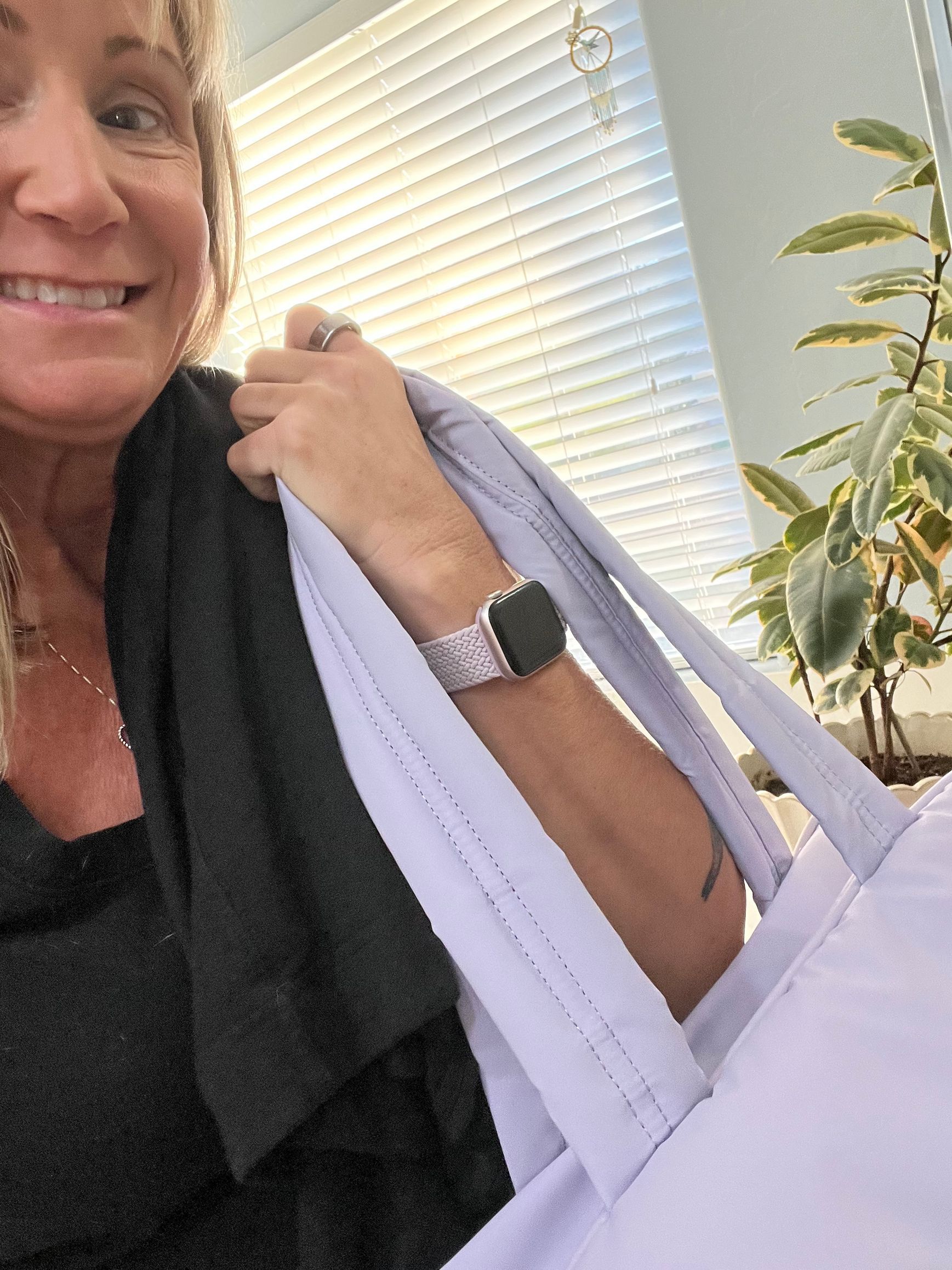 Woman smiling, carrying a lavender tote bag and a black garment over her shoulder. Indoors with a window.