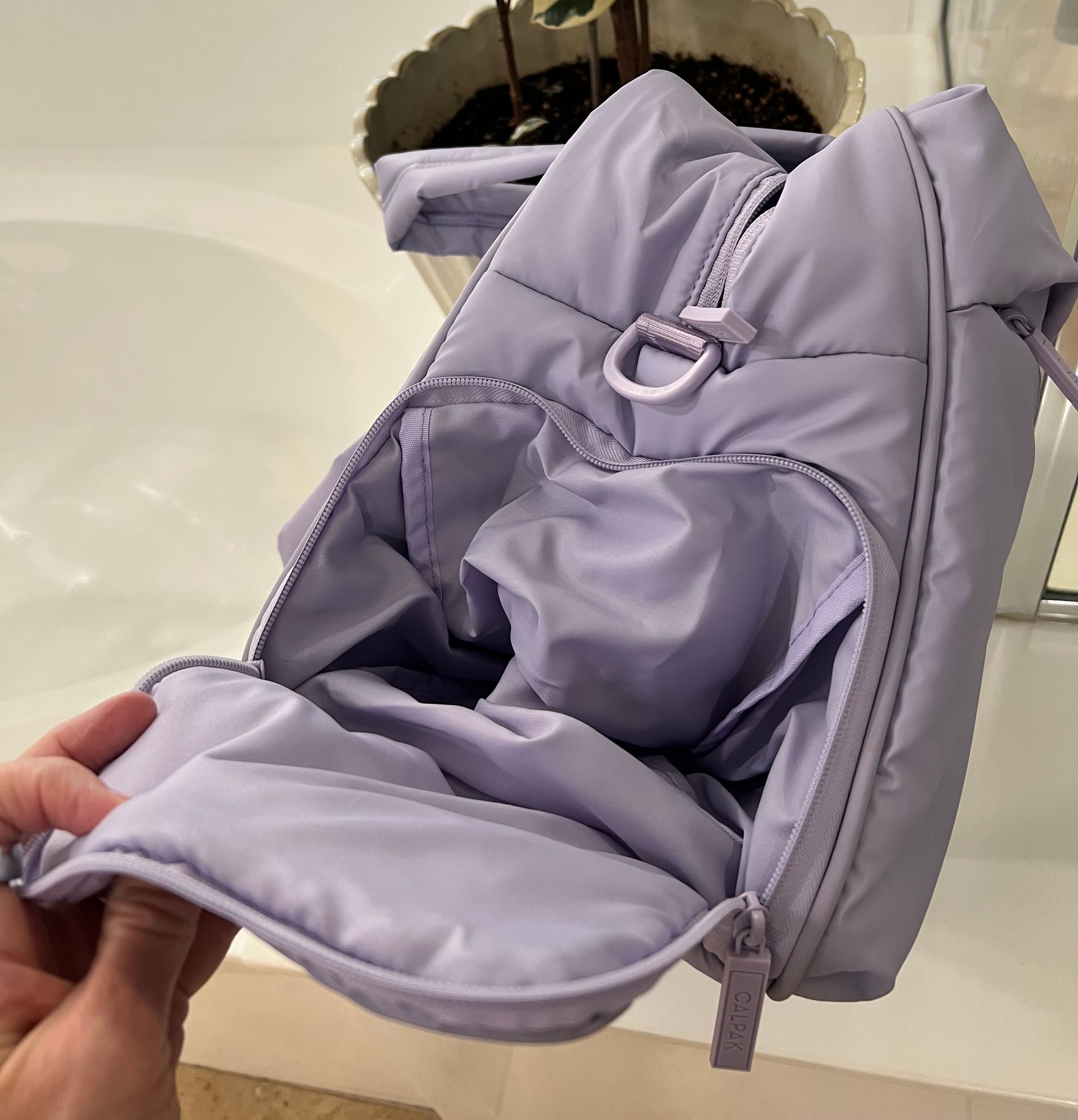 Open, light purple quilted bag with shoe compartment for travel, interior visible. A plant is in the background.
