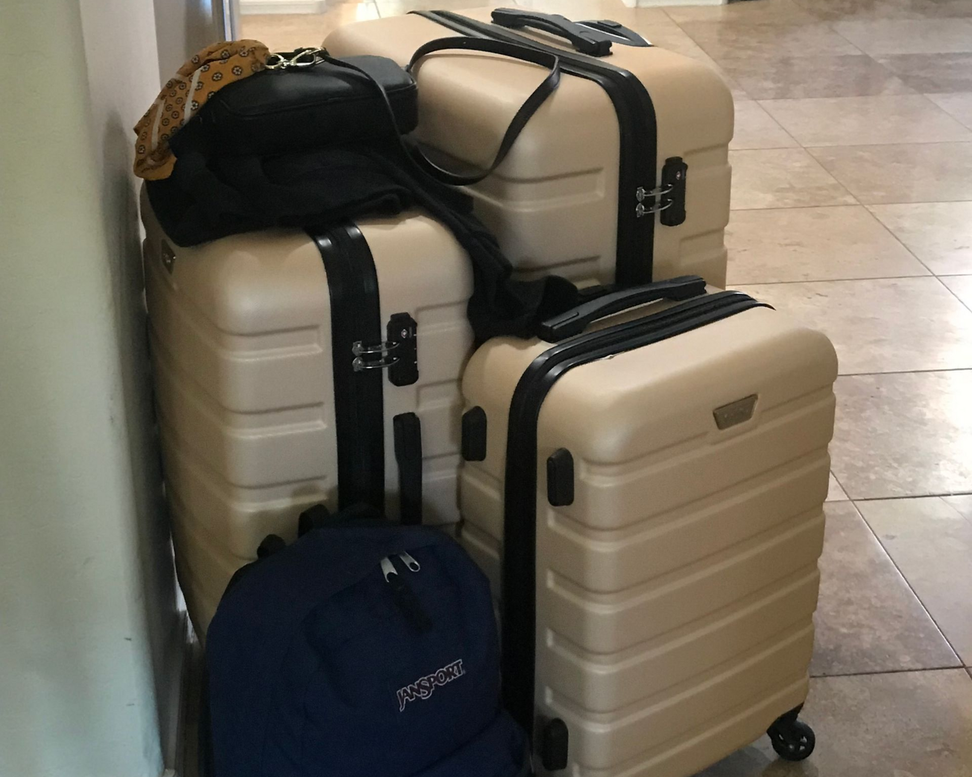 Three beige suitcases and a black backpack leaning against a wall. A black purse rests atop two suitcases.