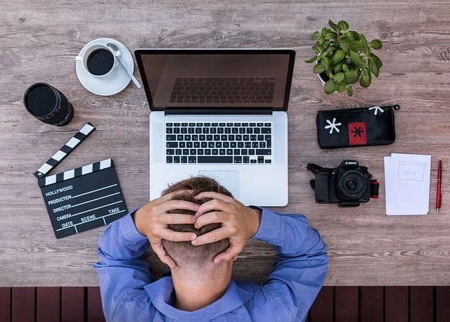 Man stressed at desk, laptop, camera, coffee, clapperboard, notepad, and plant.