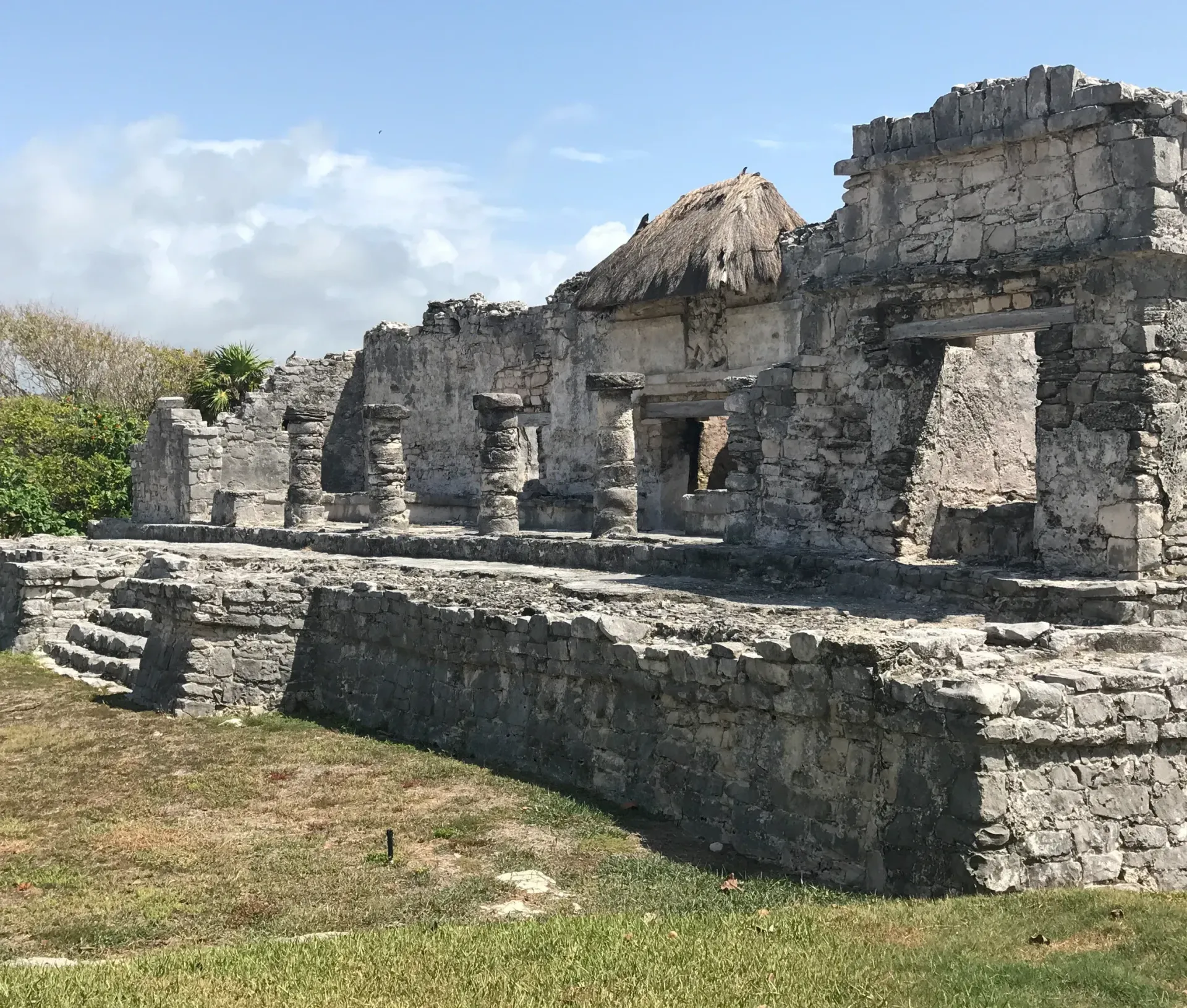 Ruined Mayan temple made of stone against a partly cloudy sky. Grass in the foreground.
