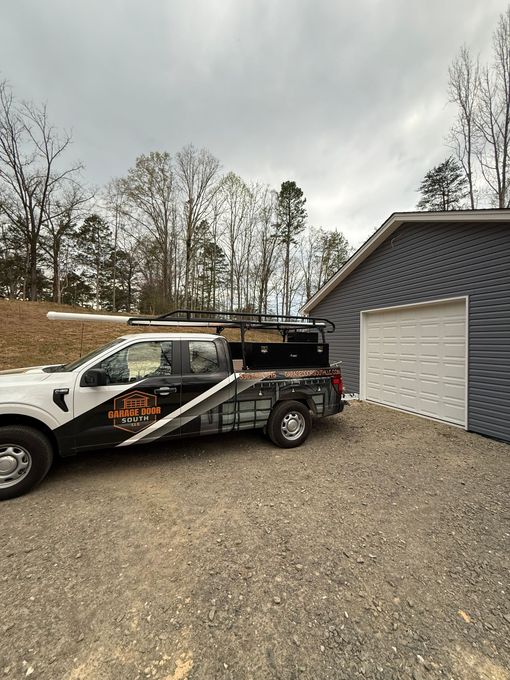 White pickup truck with graphic wrap parked beside a gray garage on a gravel driveway under a cloudy sky