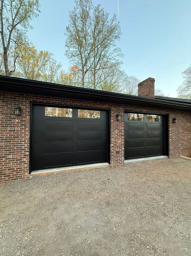 Brick garage with two black doors and a covered roof, set on a gravel driveway with trees behind it