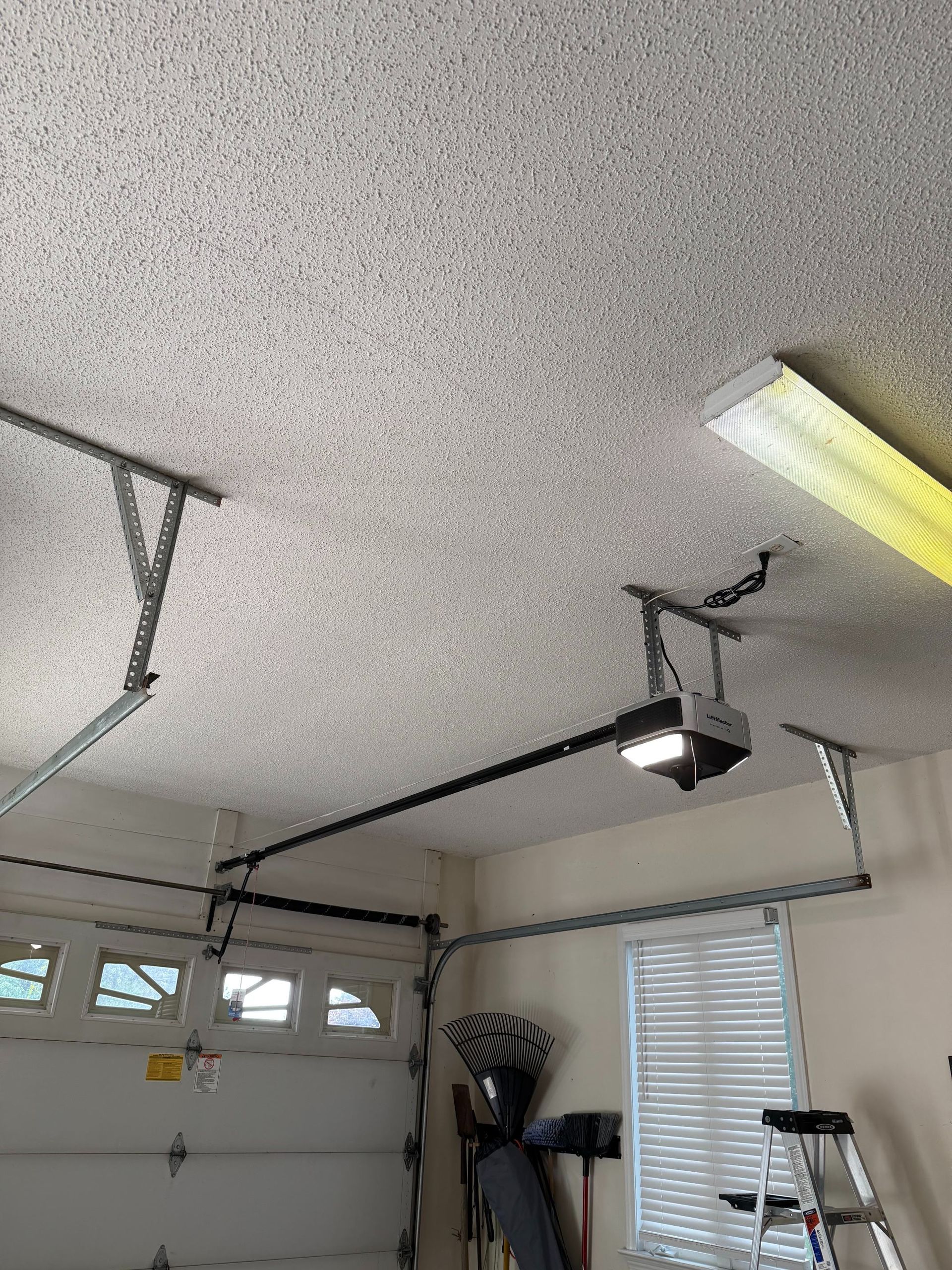 Cluttered garage with garage door, hanging opener, window, tools, and a yellow lawn tractor.