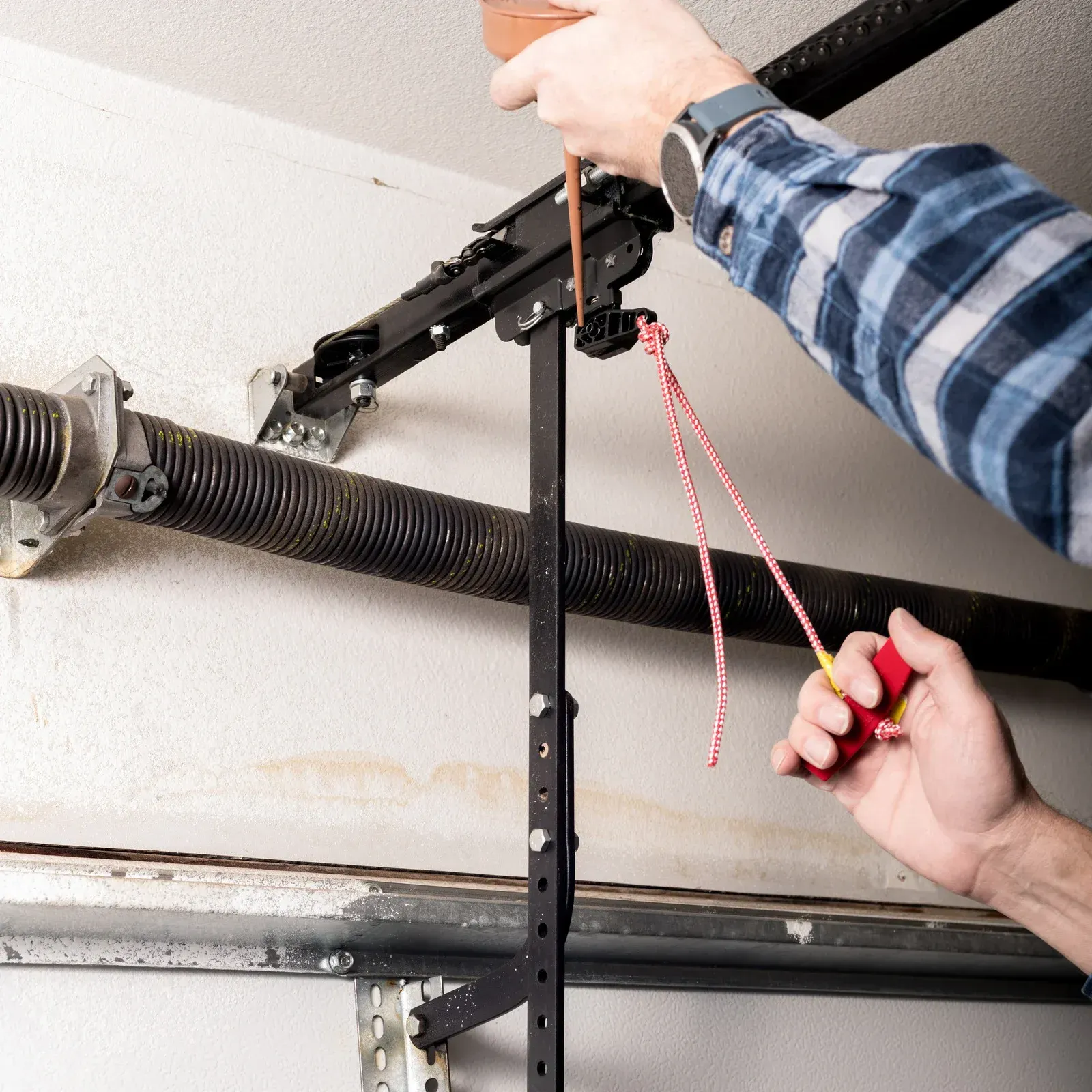 Person using a red-handled tool to adjust a black garage door spring mechanism