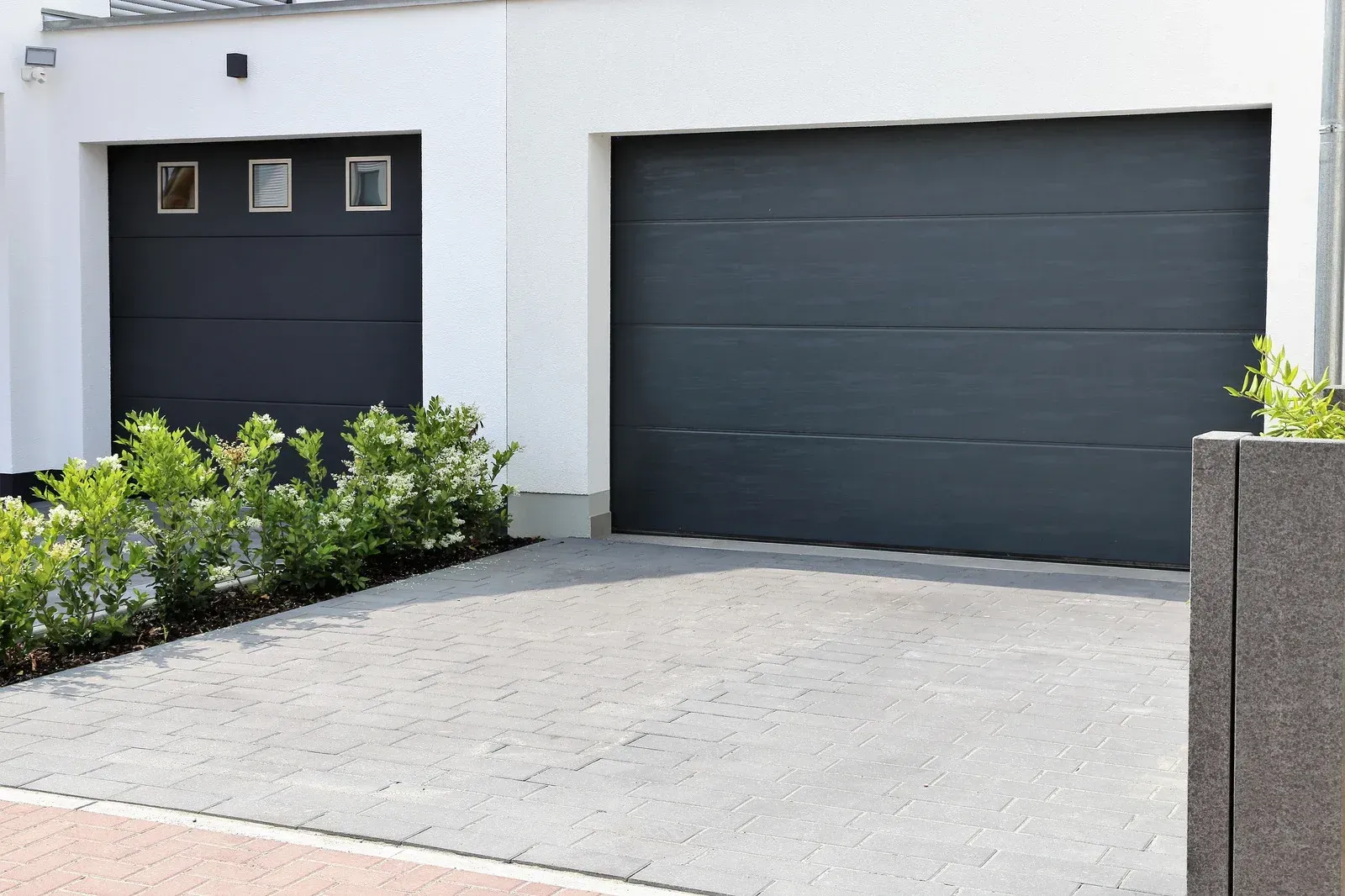 Modern house exterior with two dark garage doors and a paved driveway bordered by greenery