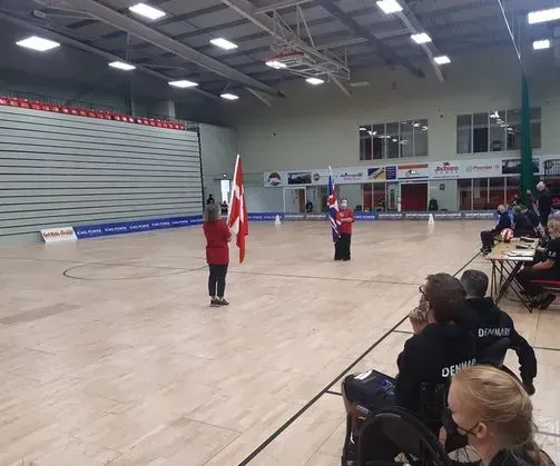 To personer holder flag i en indendørs sportshal, tilskuere siddende.