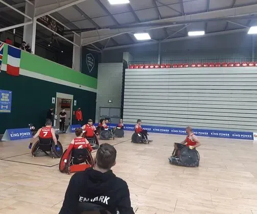 Kørestolsrugbykamp i gang indendørs. Spillere i røde og sorte trøjer konkurrerer på en bane.