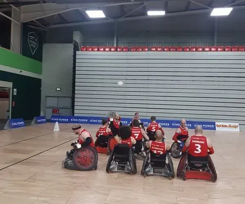 Kørestolsrugbyspillere i aktion, en med bolden, indendørsbane, røde og sorte uniformer.