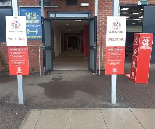 Indgang til en bygning med åbne døre, flankeret af velkomstskilte og en rød informationstavle.