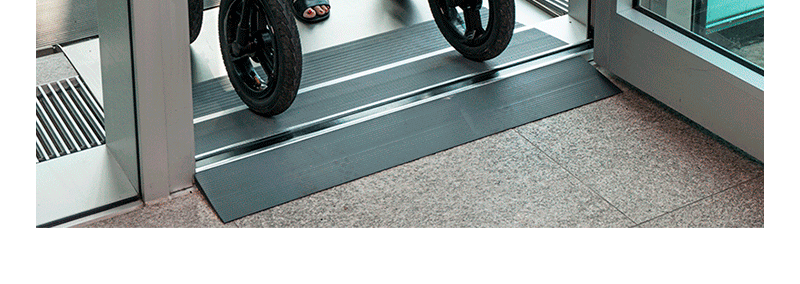 Kørestolsrampe ved en døråbning. Sorte hjul på rampen, mørkegrå rampe, lyst tæppe på gulvet.