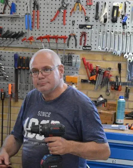 Mand holder en boremaskine i et værksted, værktøj på væggen bag sig.