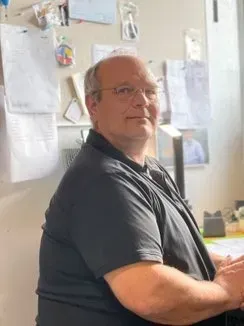 Mand med briller og sort skjorte, siddende ved et skrivebord, kigger mod kameraet, kontoromgivelse.