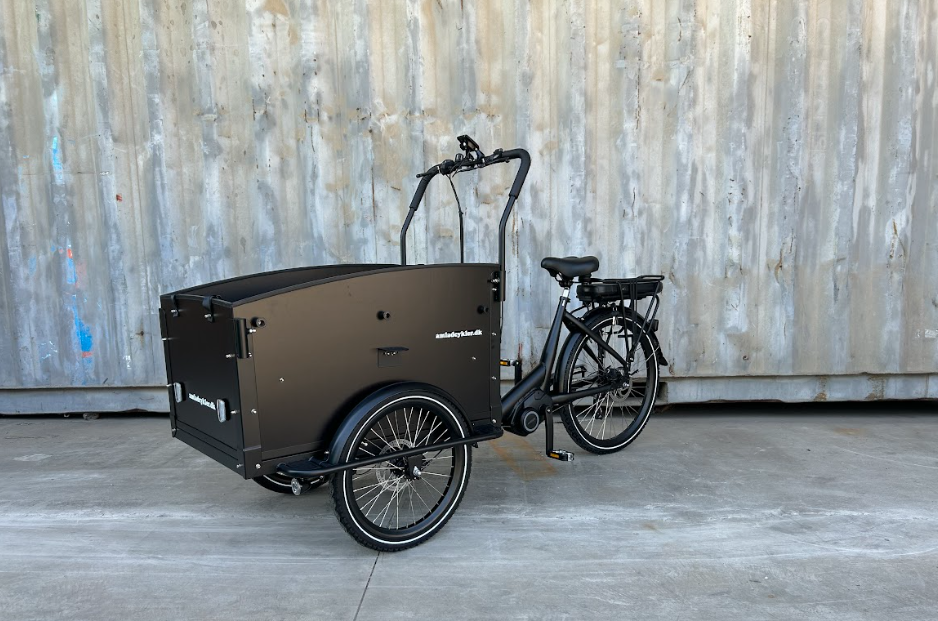 Sort ladcykel med stor frontboks, parkeret foran en struktureret væg.