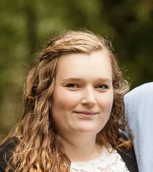 Kvinde med lys hud og krøllet blond hår smiler til kameraet, iført en sort cardigan og hvid blondetop, udendørs.